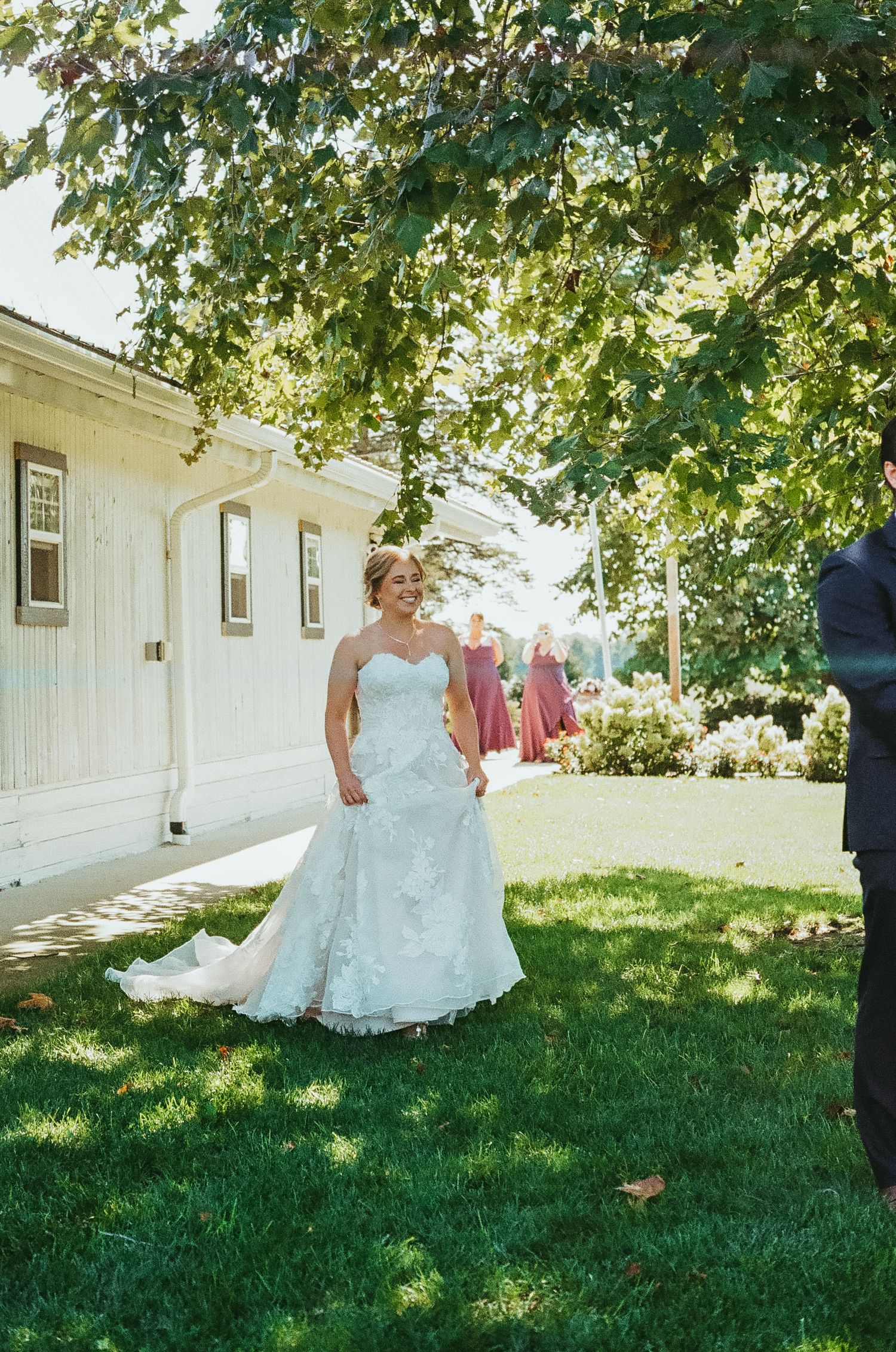 Smiling bride in strapless lace wedding gown with train standing on green lawn outside white building on sunny day.