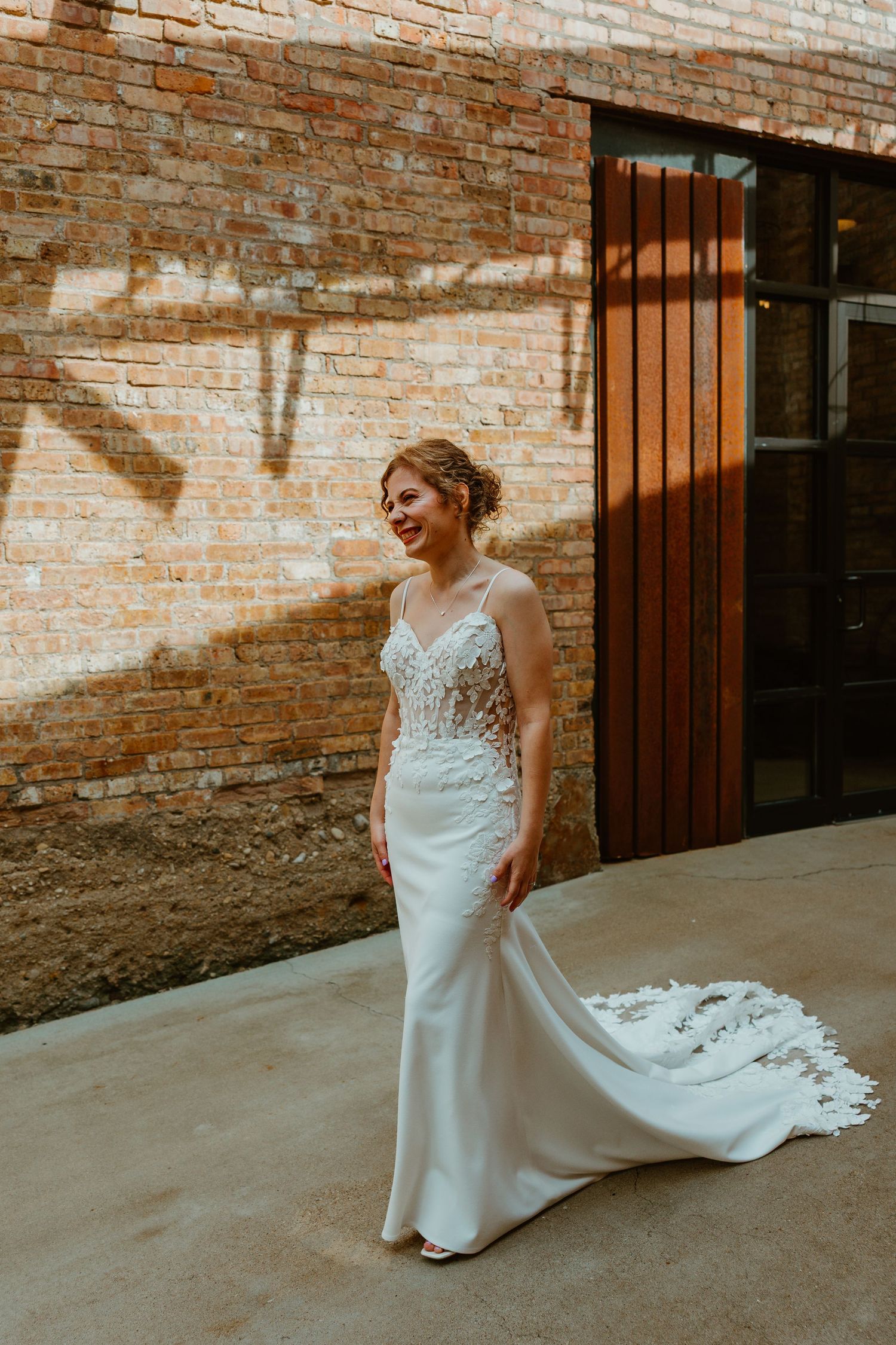 Bride in a fitted lace wedding gown posing outdoors against a sunlit rustic brick wall. Chicagoland wedding venues.