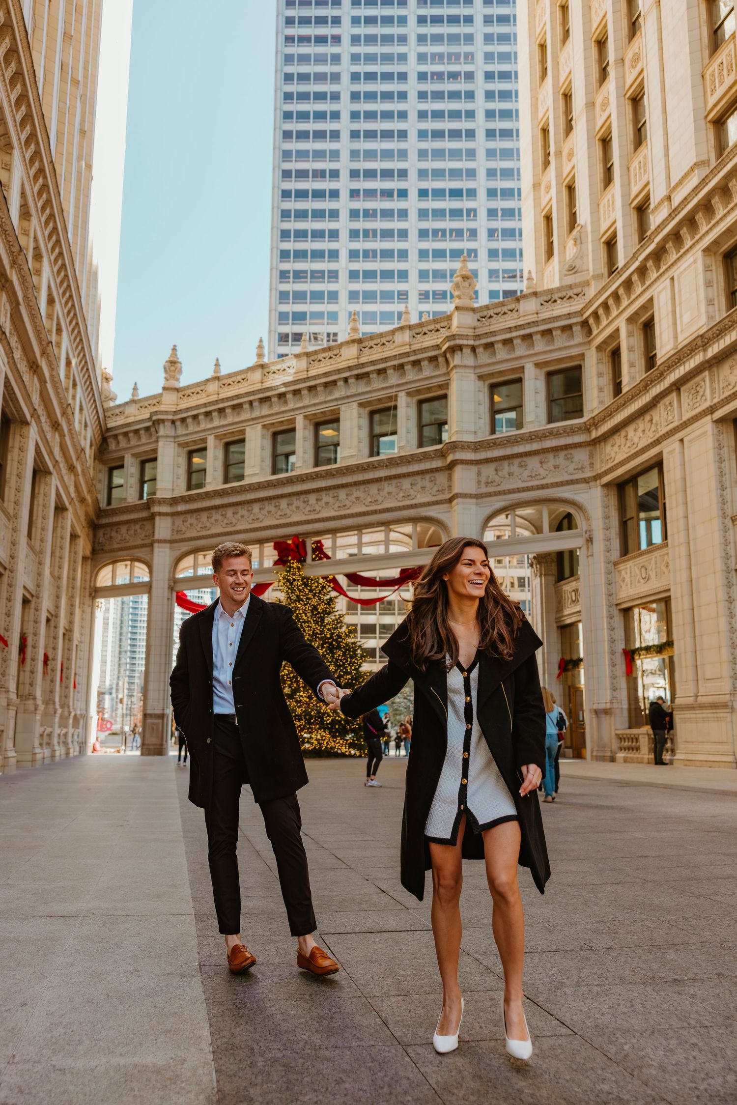 Happy couple walking hand in hand through a grand urban arcade with holiday decorations. Outdoor Chicago engagement photos.