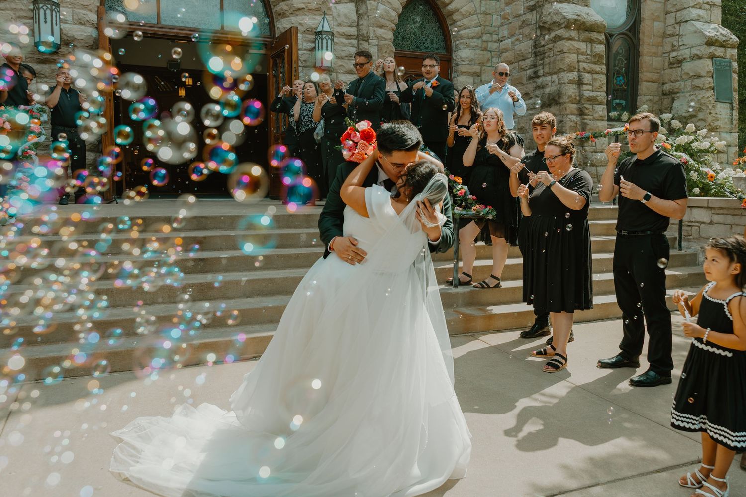 Newlyweds share a romantic kiss surrounded by bubbles outside a church after ceremony. Catholic ceremony in Chicago Catholic Church.