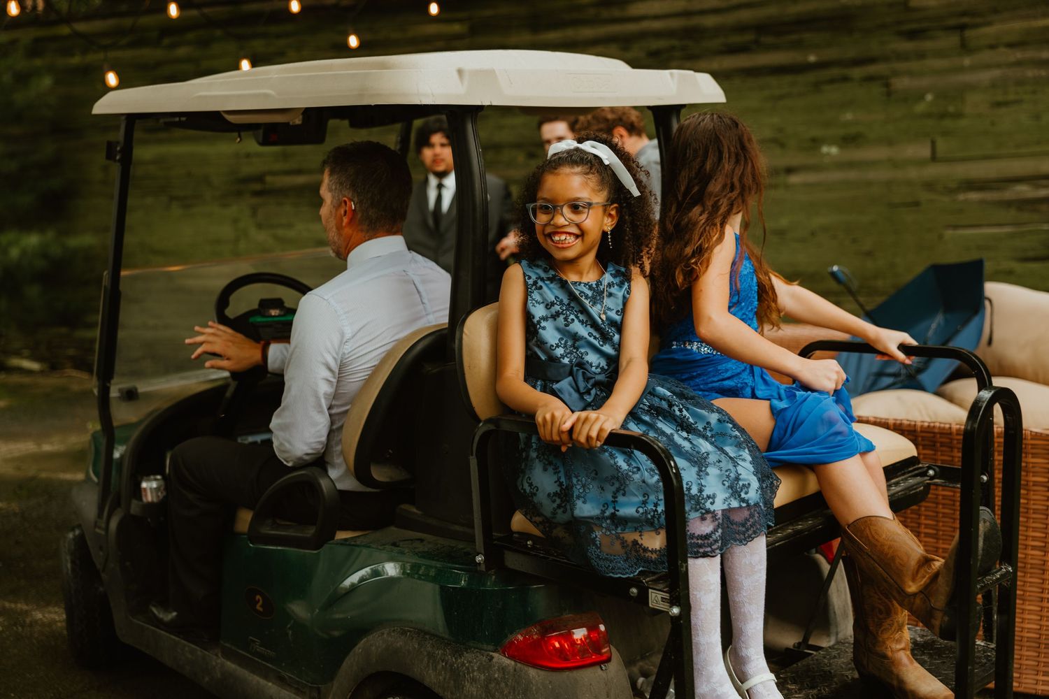 Children in blue floral dresses smile and ride golf cart at outdoor wedding reception at dusk. Northwest Indiana wedding venues.