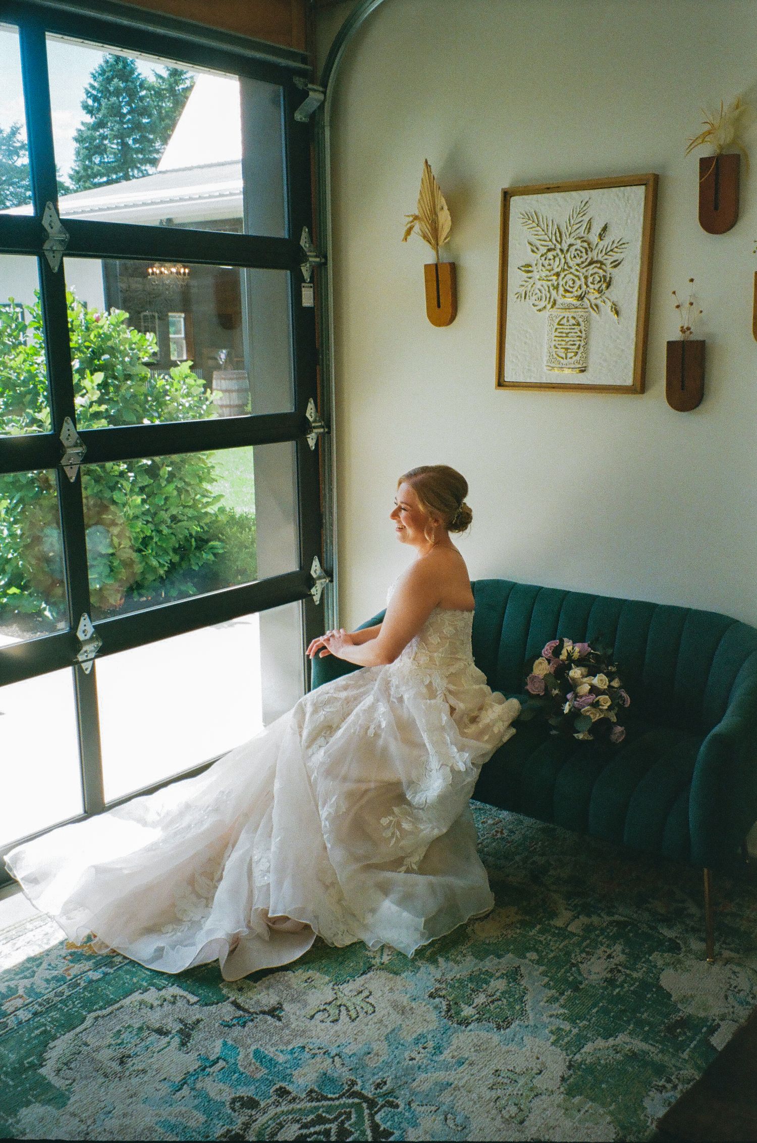 Bride in a white lace gown sits on a teal velvet sofa gazing out a large window, holding a bouquet.