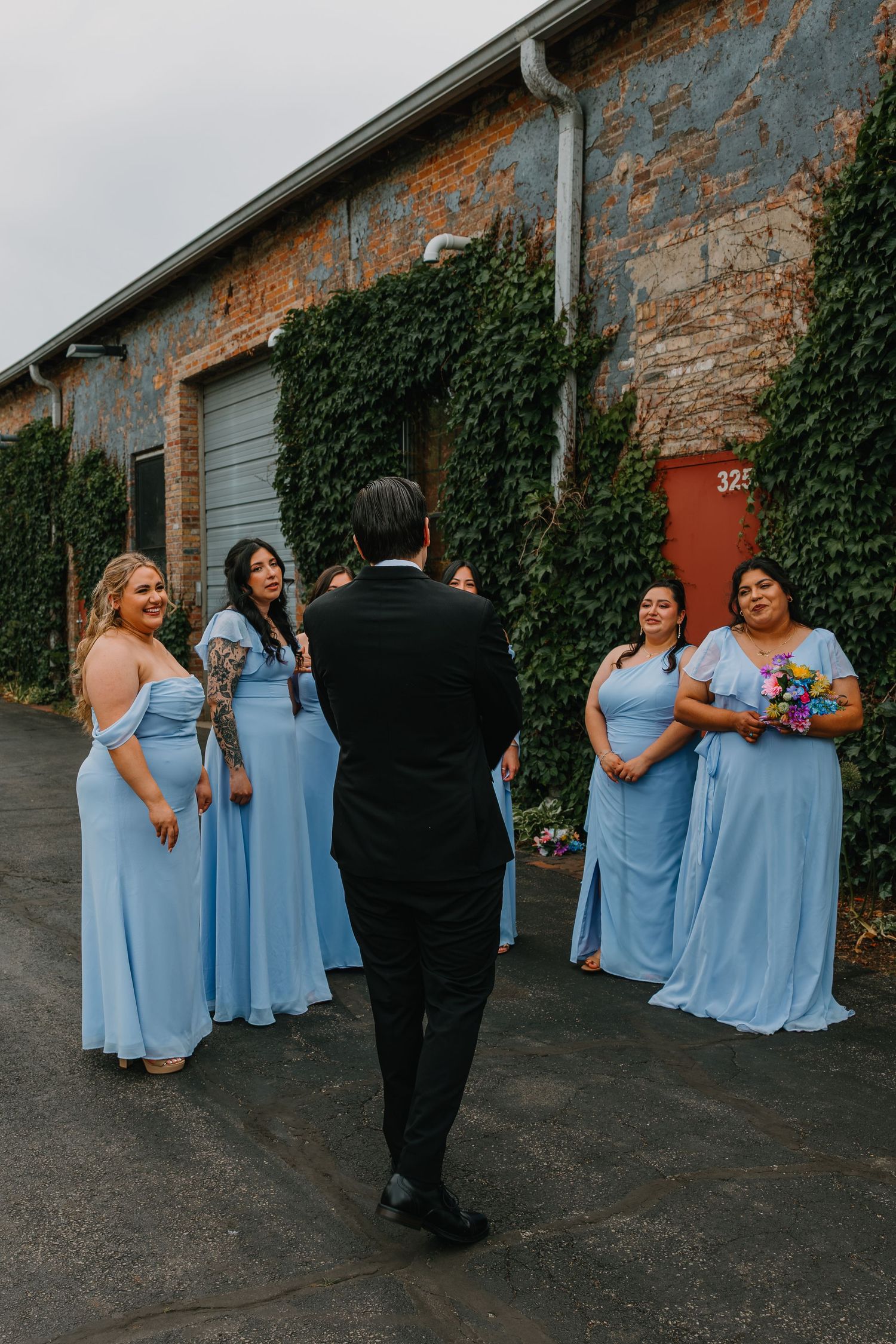 Four bridesmaids in light blue gowns smile as groom in black tux approaches them outside brick building. Chicagoland wedding venues.