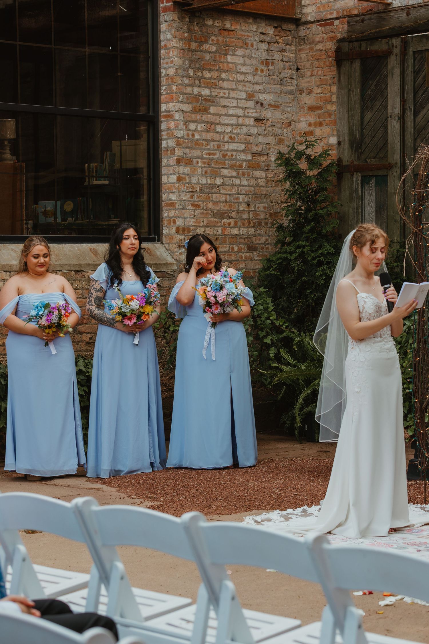 Bride reads vows at microphone while emotional bridesmaids in blue dresses stand beside her at outdoor ceremony. Chicagoland wedding venues.