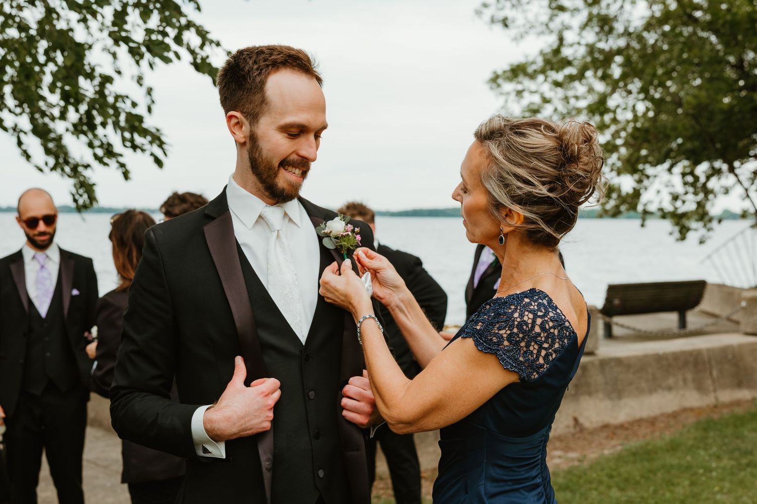 Mother in navy lace dress pinning boutonniere on groom in black tuxedo near lakeside venue.