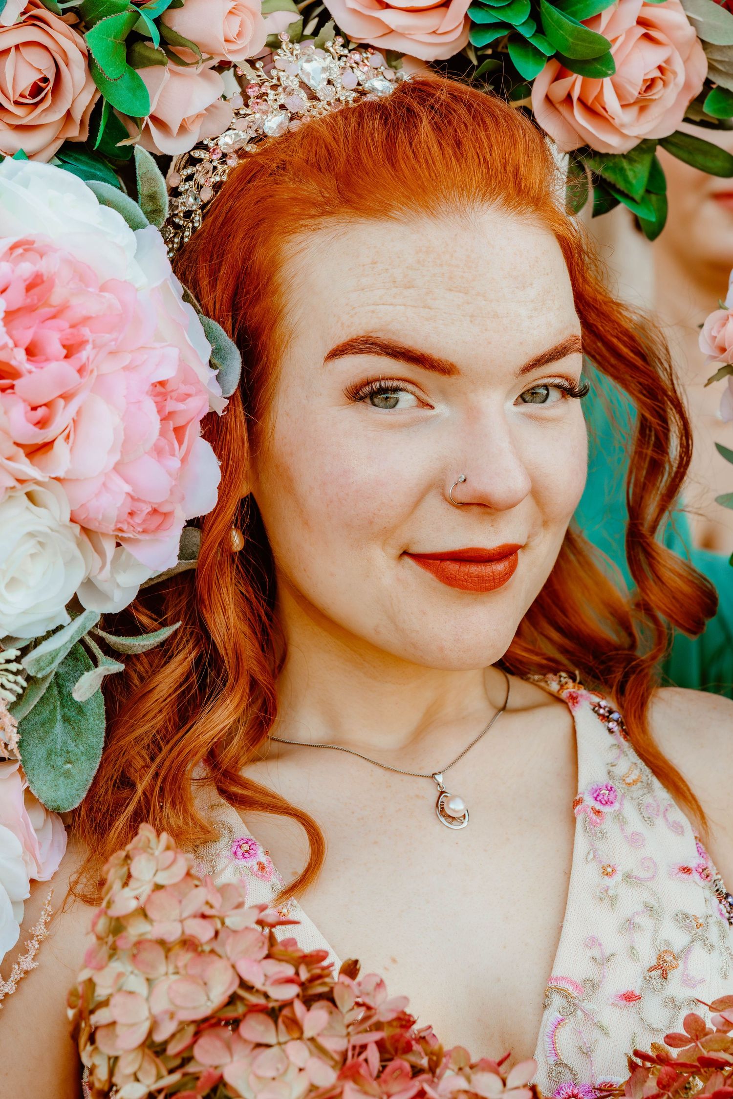 Close-up portrait of a freckled red-haired bride with tiara surrounded by pink flowers.