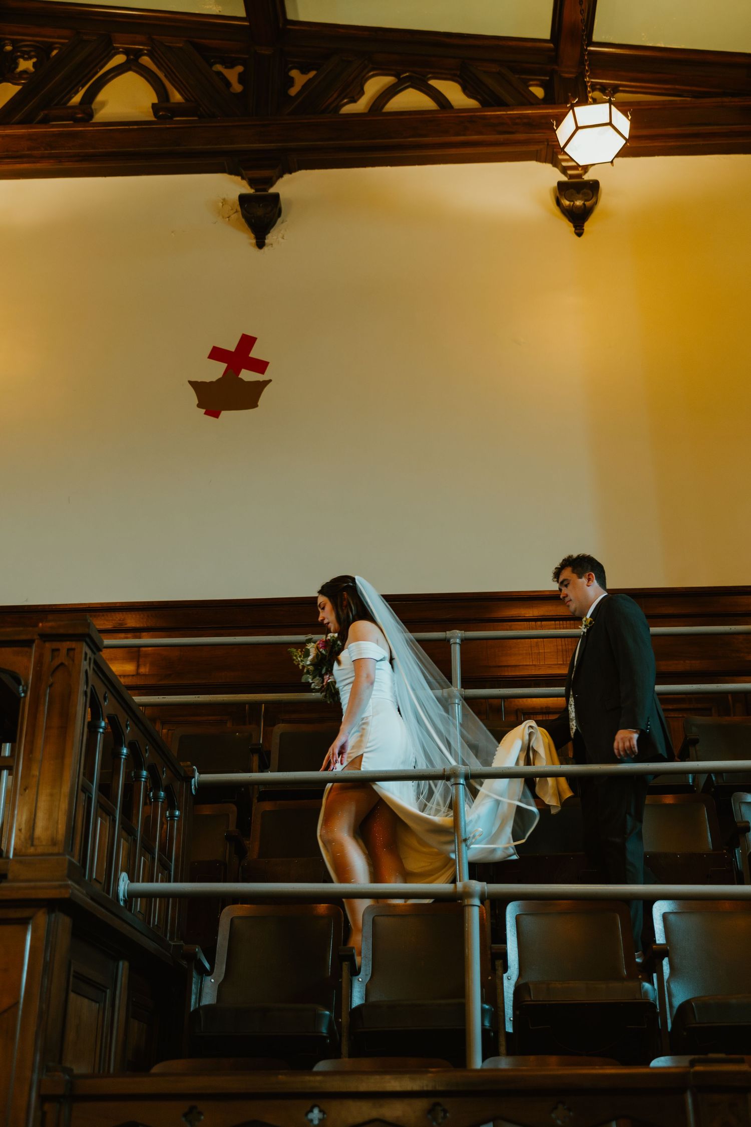 Bride and groom walking down stairs in a historic church with wooden pews and a red cross on the wall.