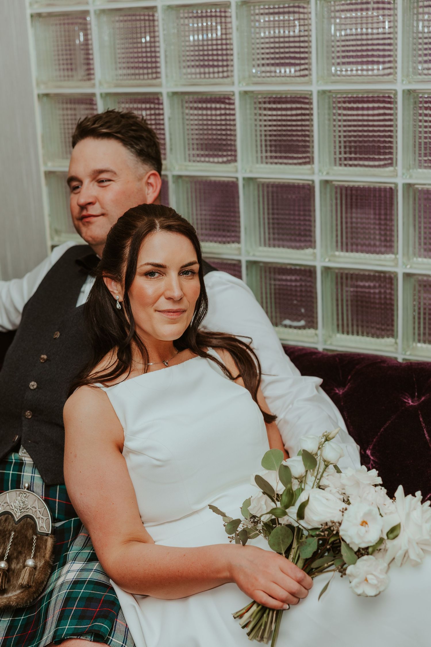 Bride holding white bouquet sits with groom in tartan kilt on sofa. Chicago Thompson Hotel wedding.