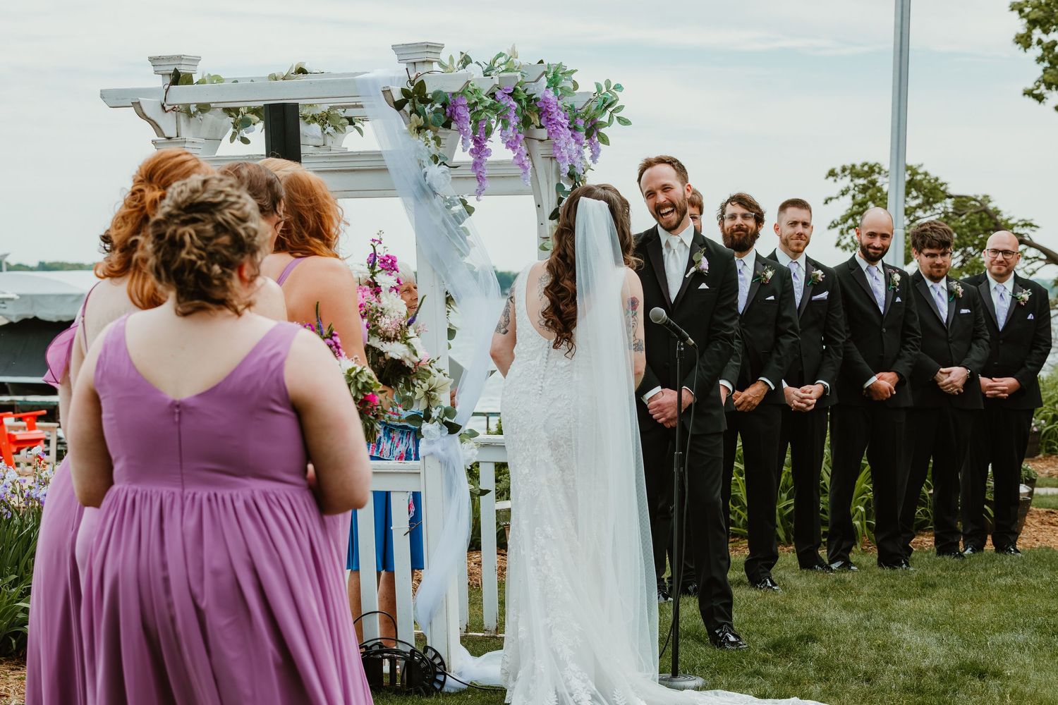 Groom smiling as bride approaches the outdoor wedding altar decorated with purple floral arch.