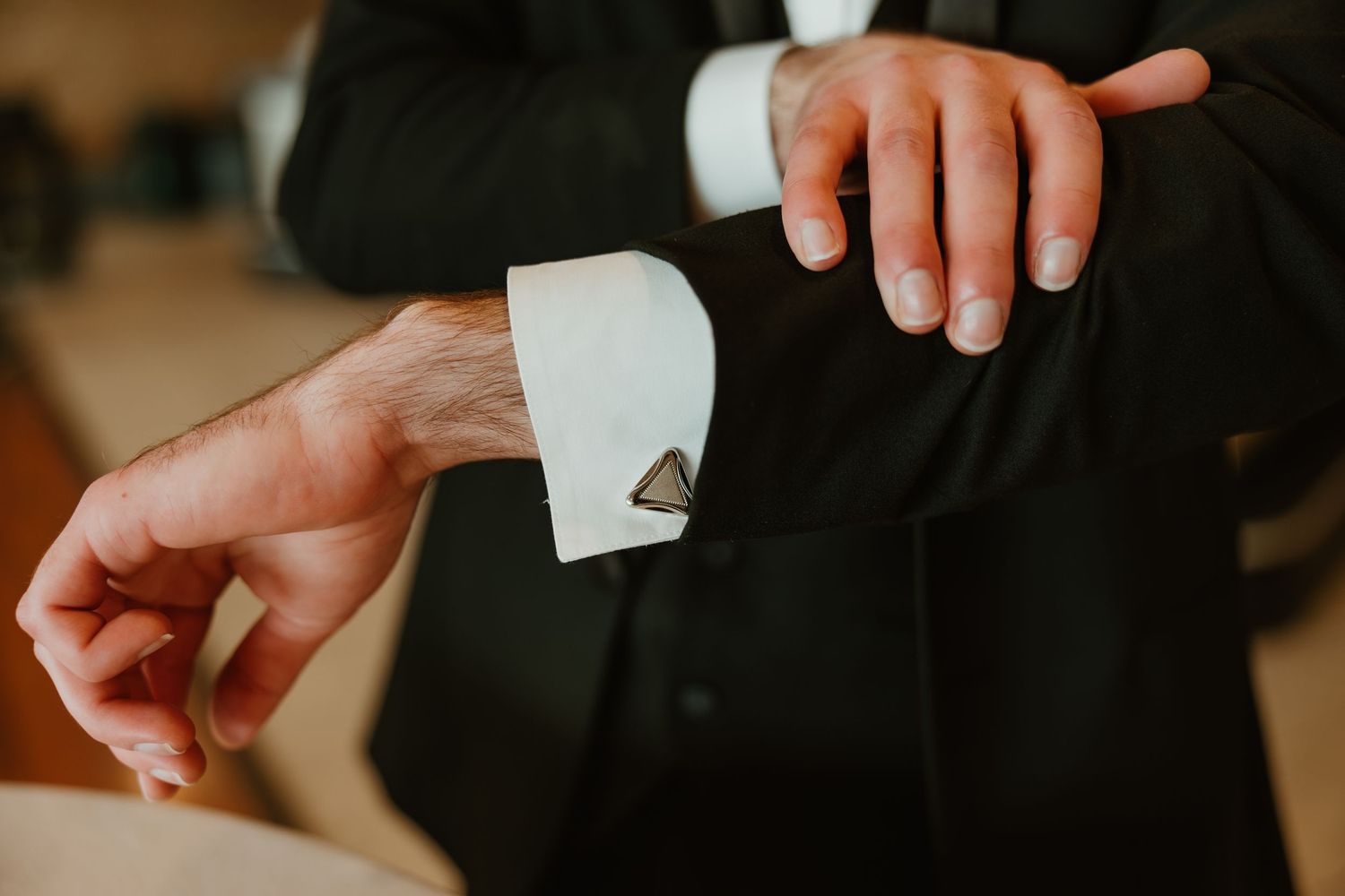 Close-up of groom fastening triangular cufflinks on white dress shirt cuff of black tuxedo.