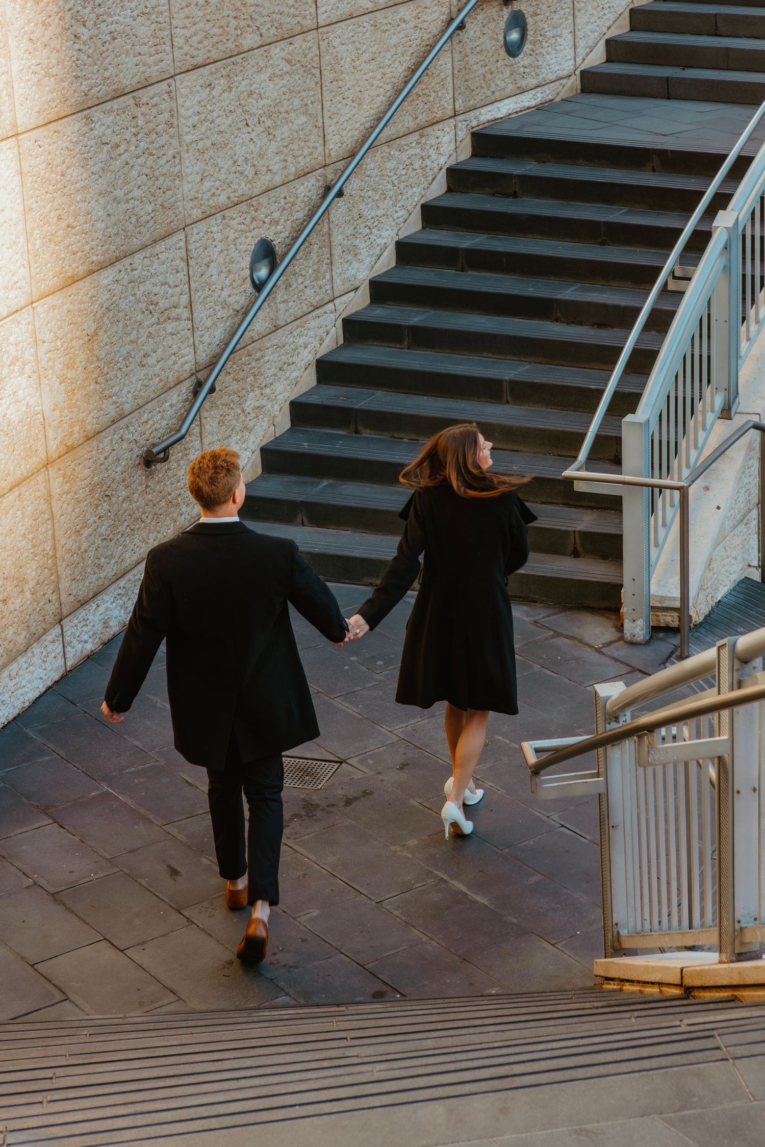 Couple holding hands walking up outdoor urban stairs in matching black coats. Outdoor Chicago engagement photos.
