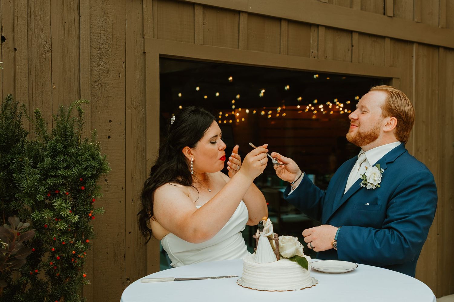 Bride and groom feeding each other wedding cake at their outdoor barn reception with string lights. Northwest Indiana wedding venues.