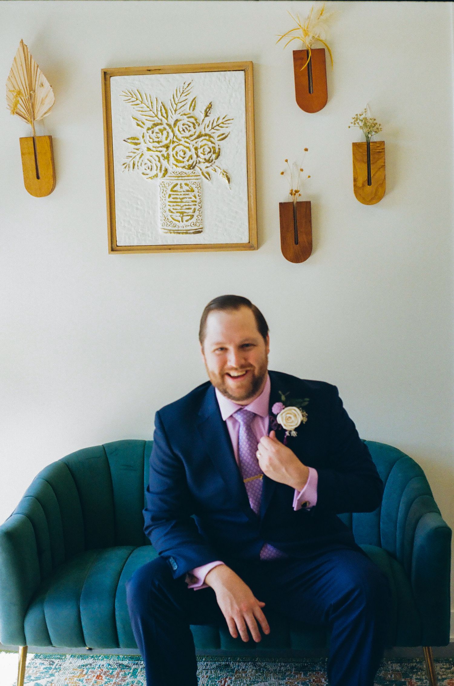 Smiling groom in navy suit with boutonniere sits on teal velvet sofa beneath floral wall art at wedding venue.