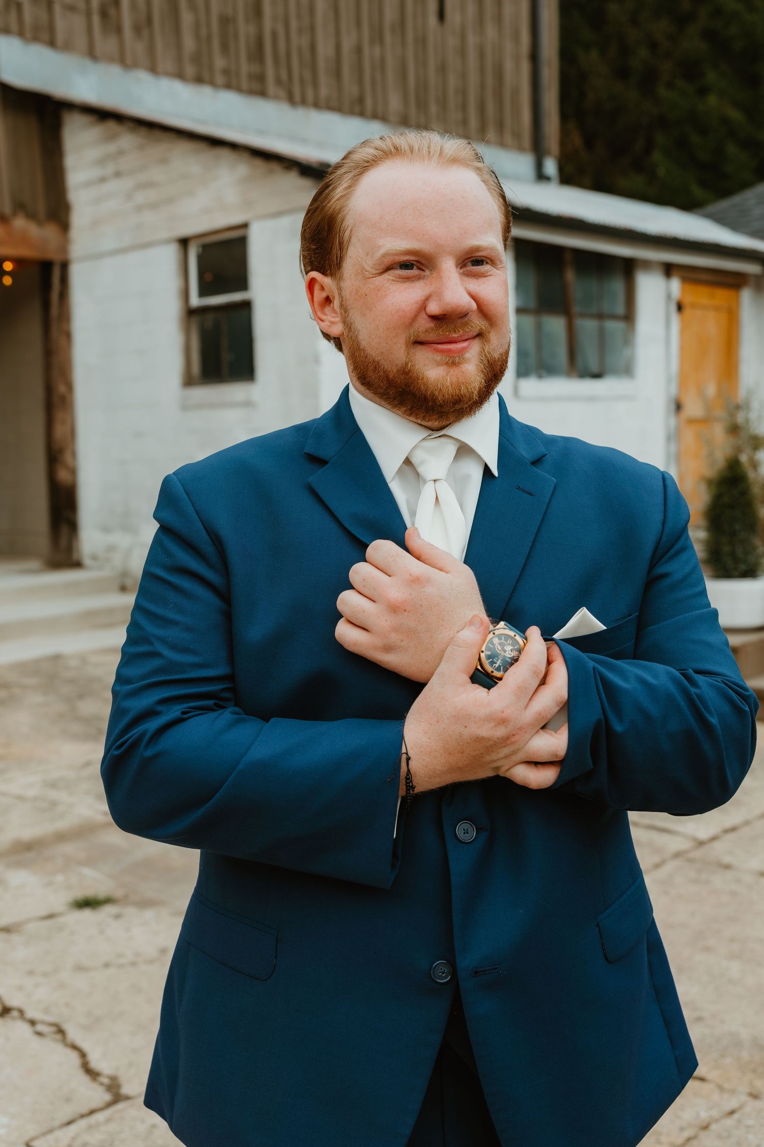Handsome groom in a navy blue suit adjusts his cufflinks while smiling outside a rustic barn wedding venue. Northwest Indiana wedding venues