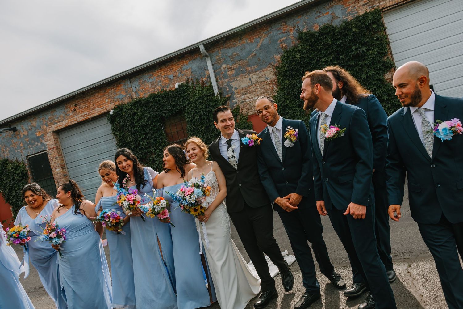 Wedding party in light blue and navy attire walks together laughing outside ivy-covered brick venue. Chicagoland wedding venues.