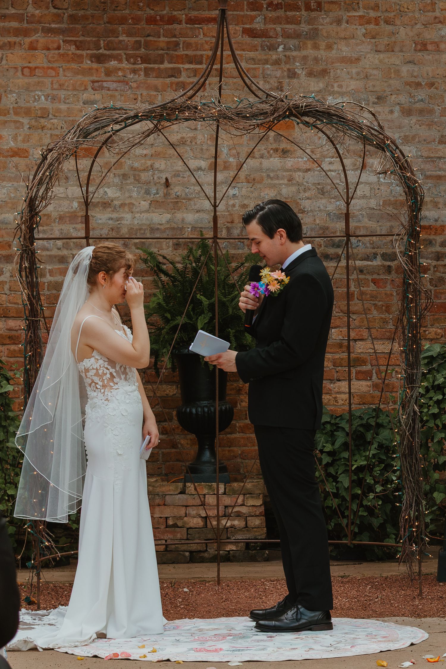 Bride wipes tears as groom reads heartfelt vows under rustic wire arch at outdoor brick wall ceremony. Chicagoland wedding venues.