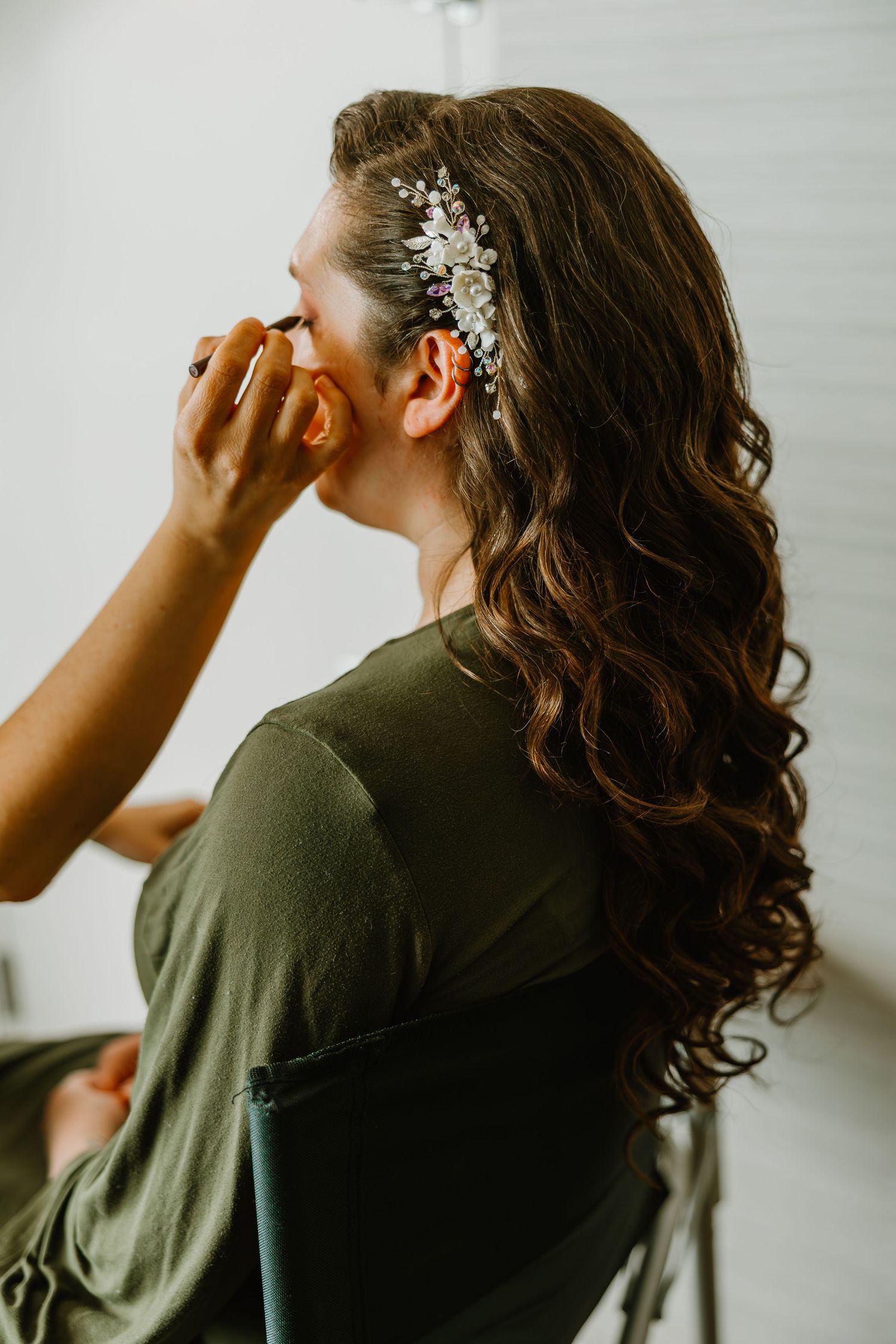 Bride with long curly hair and floral hair comb getting makeup applied while wearing green robe.