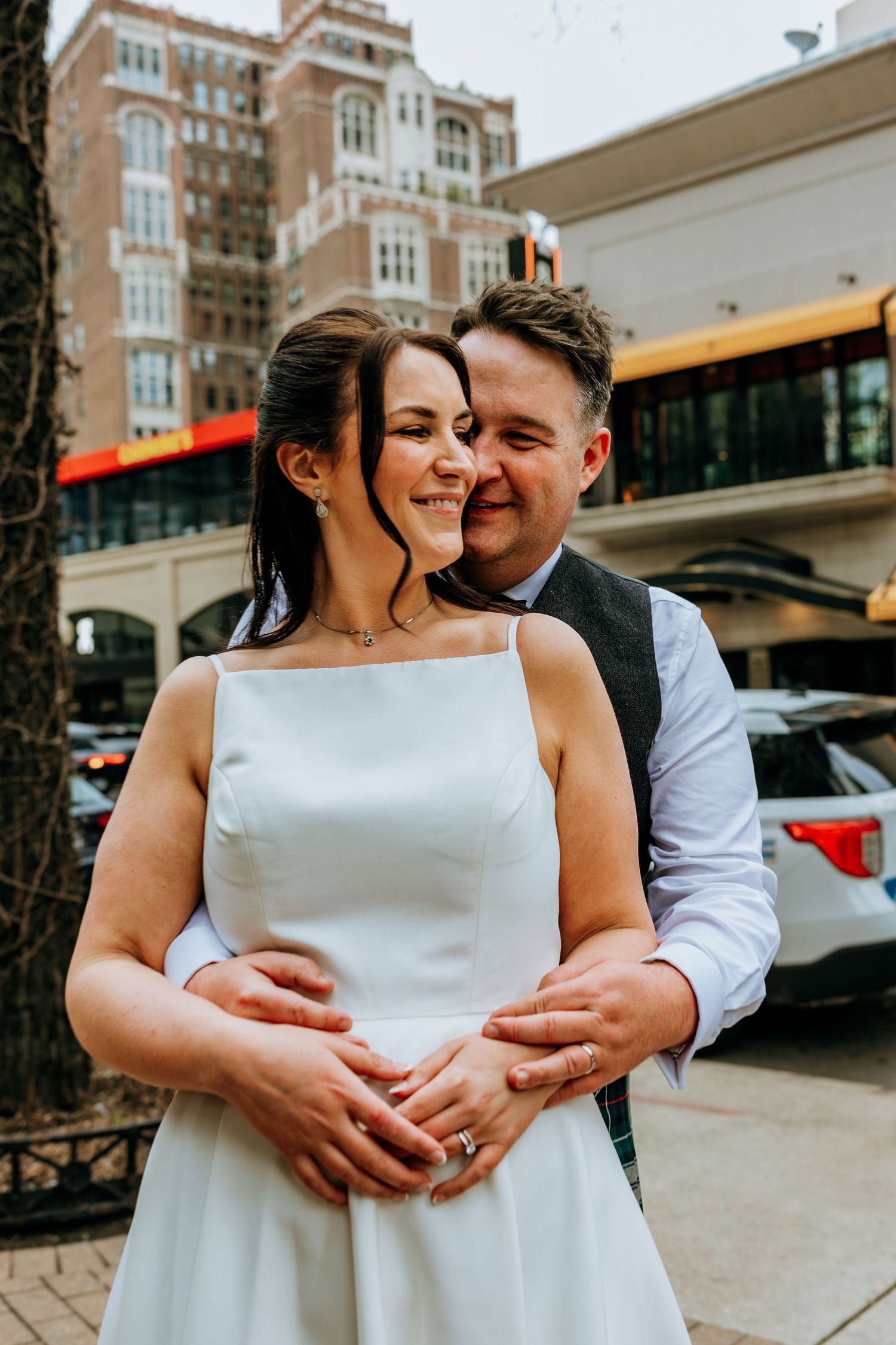 Groom embraces bride in white square-neck dress from behind on city street at their wedding. Downtown Chicago elopement.