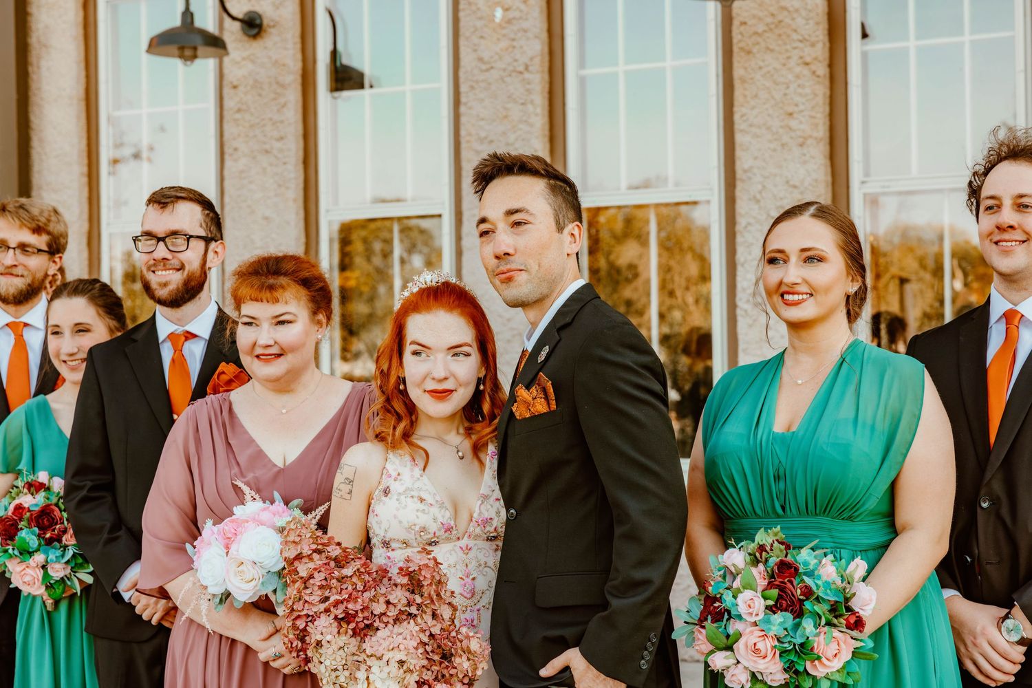 Wedding party posing outside venue with bride in floral dress and bridesmaids in teal gowns.