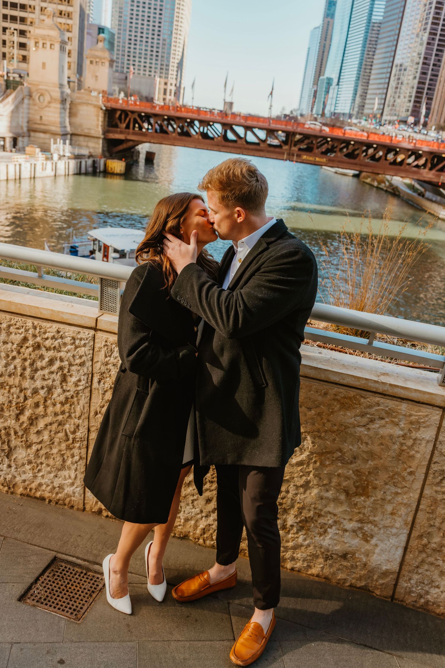 Couple kissing on a riverside walkway with a historic bridge and city skyline behind them. Outdoor Chicago engagement photos.