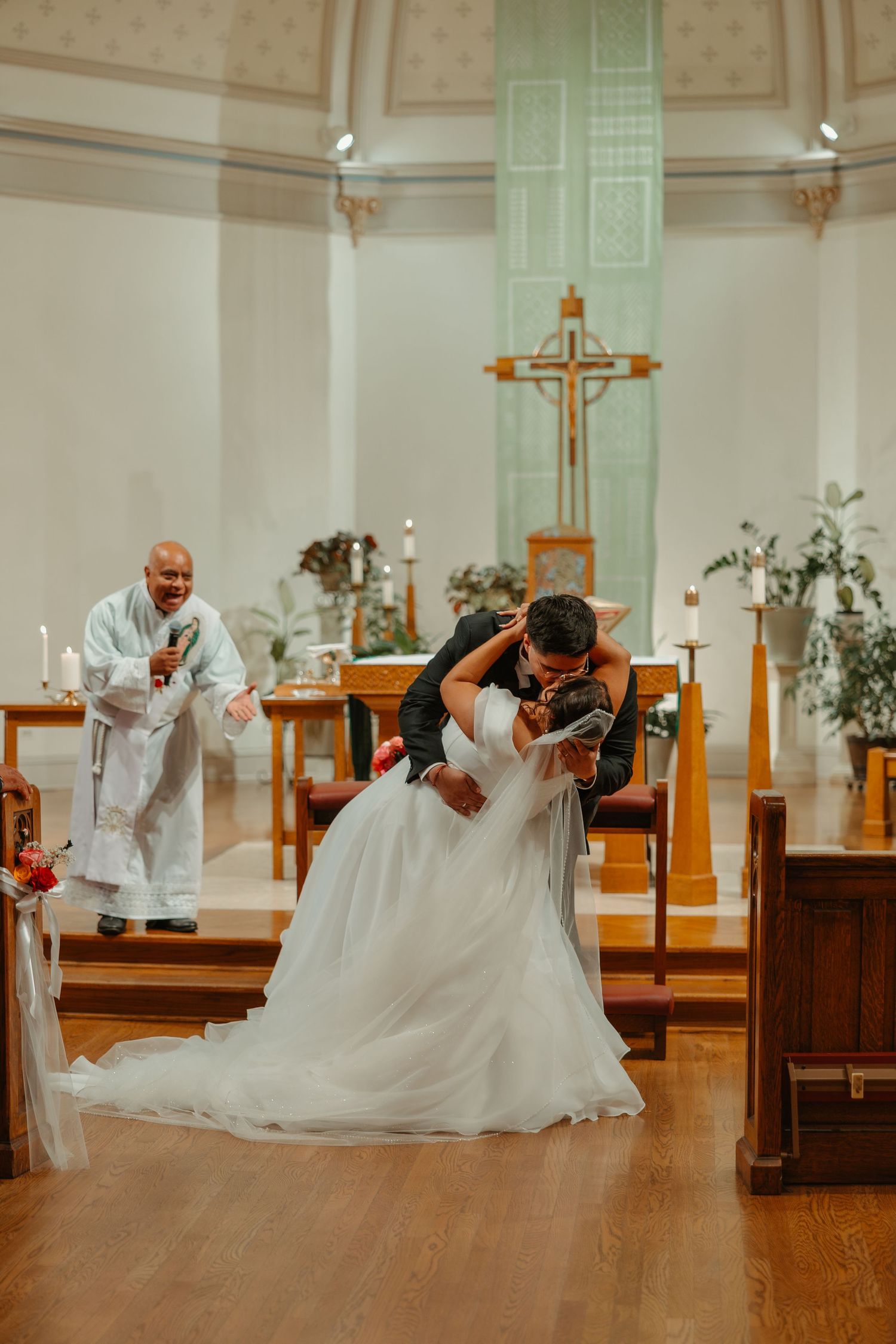 Groom dipping bride in a romantic kiss at the altar as priest celebrates behind them. Catholic ceremony in Chicago Catholic Church.
