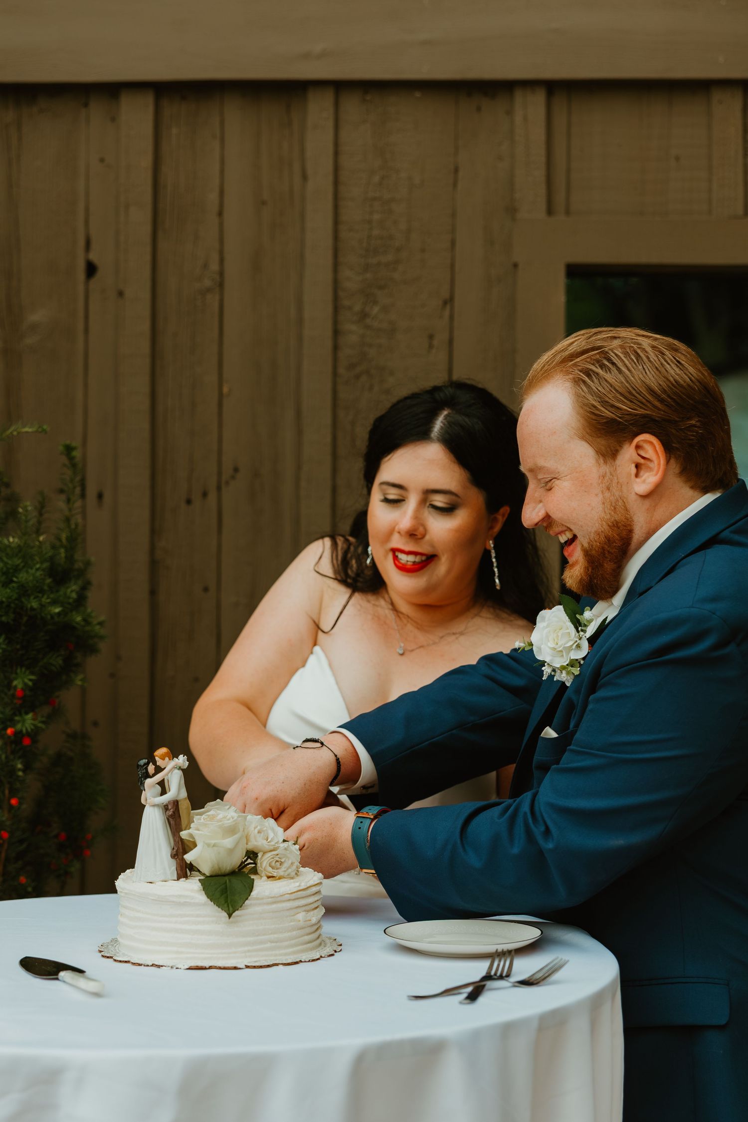 Bride and groom smiling while cutting their white wedding cake together at an outdoor barn reception. Northwest Indiana wedding venues.