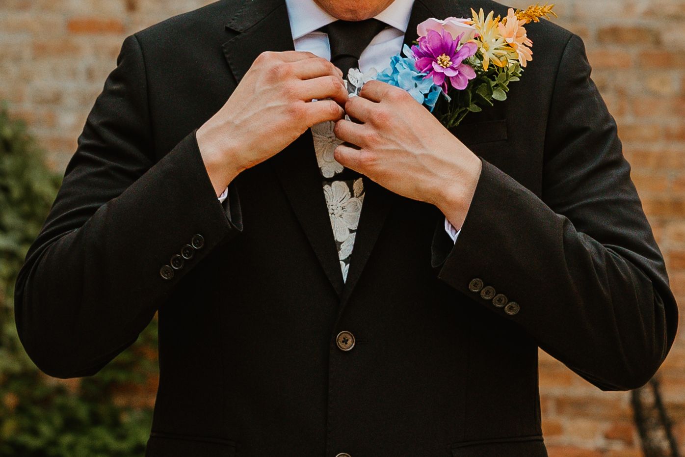 Groom in a black suit adjusting his colorful floral boutonniere against a brick background. Chicagoland wedding venues.
