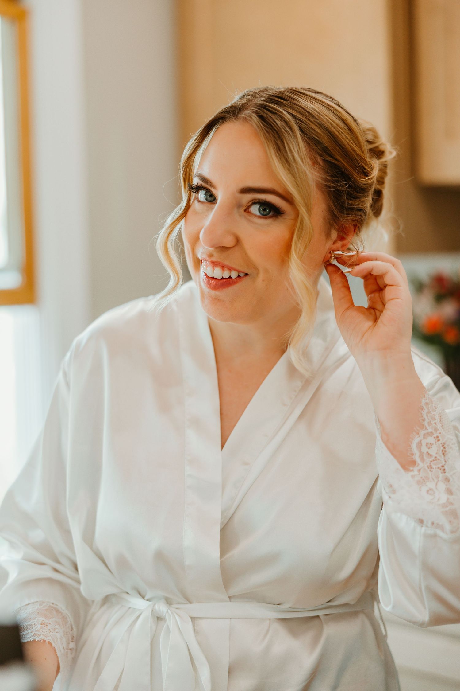 Beautiful bride in white lace robe smiling while putting on earrings during wedding day preparations. Chicago documentary style photography.