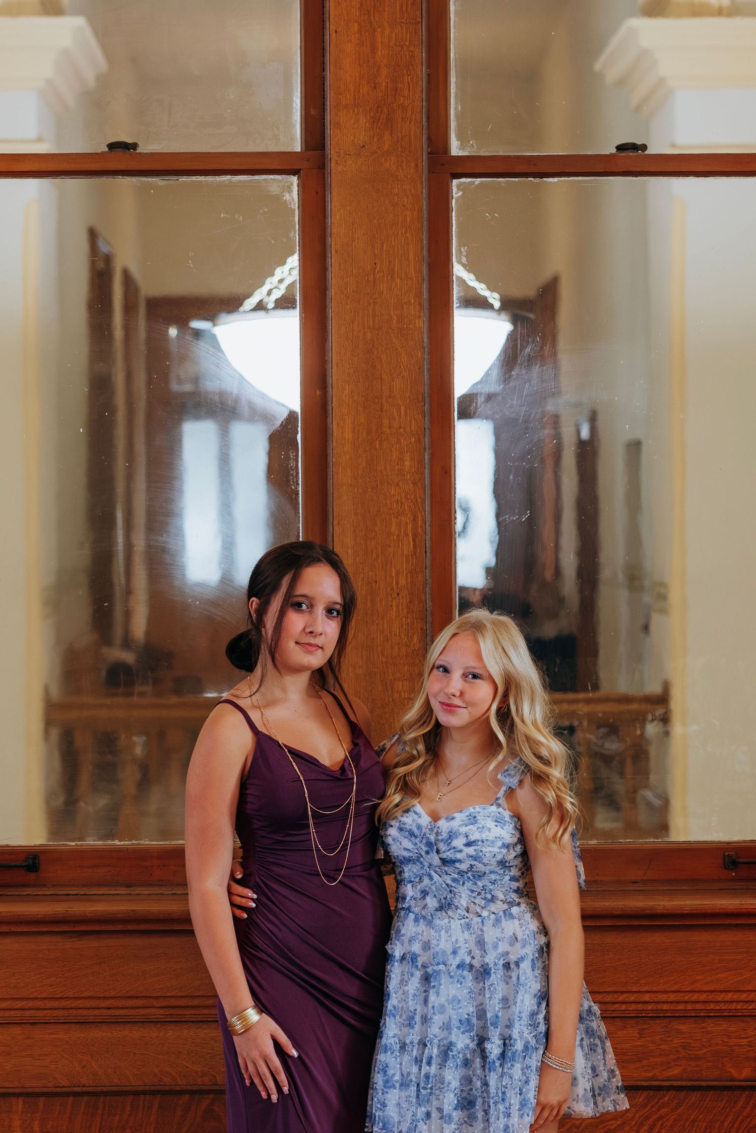 Two young women in formal dresses posing together in front of a wooden cross window at a wedding venue. Crown Point Courthouse wedding.