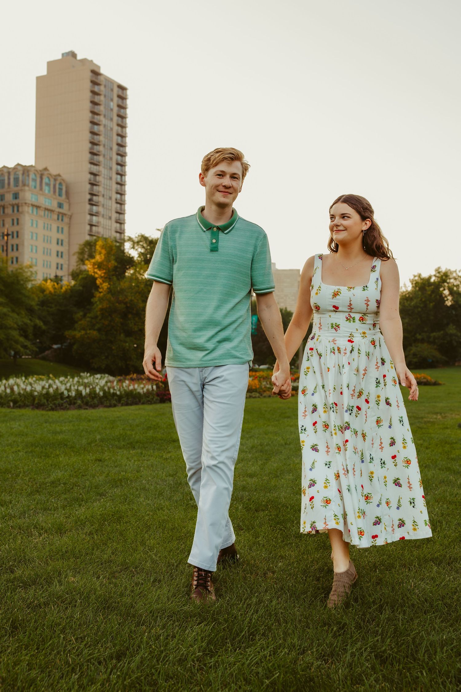 Young couple holding hands and walking on green grass in a city park with tall buildings behind them. Chicago engagement photos.