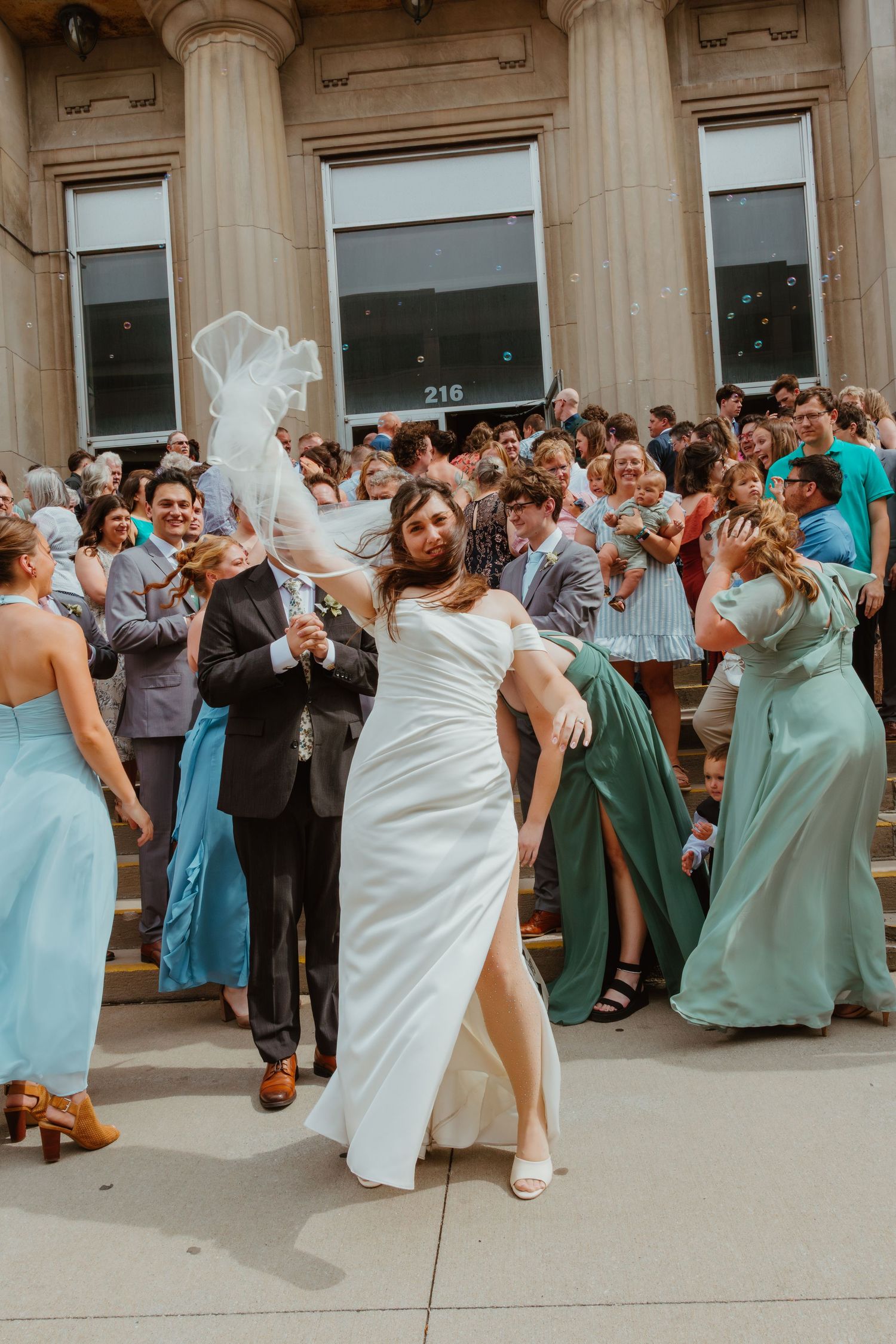 Joyful bride in white wedding dress tossing veil on steps with wedding party and guests behind her.