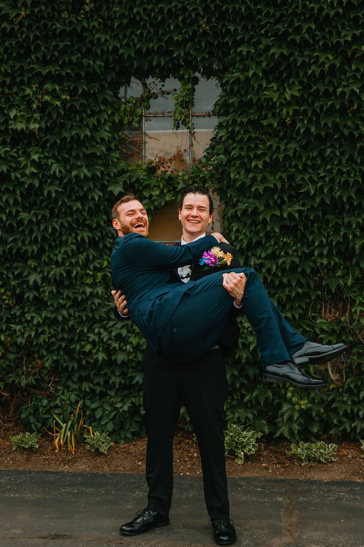 Groomsman in navy suit carries laughing groom in black tux in front of ivy-covered wall outdoors. Chicagoland wedding venues.