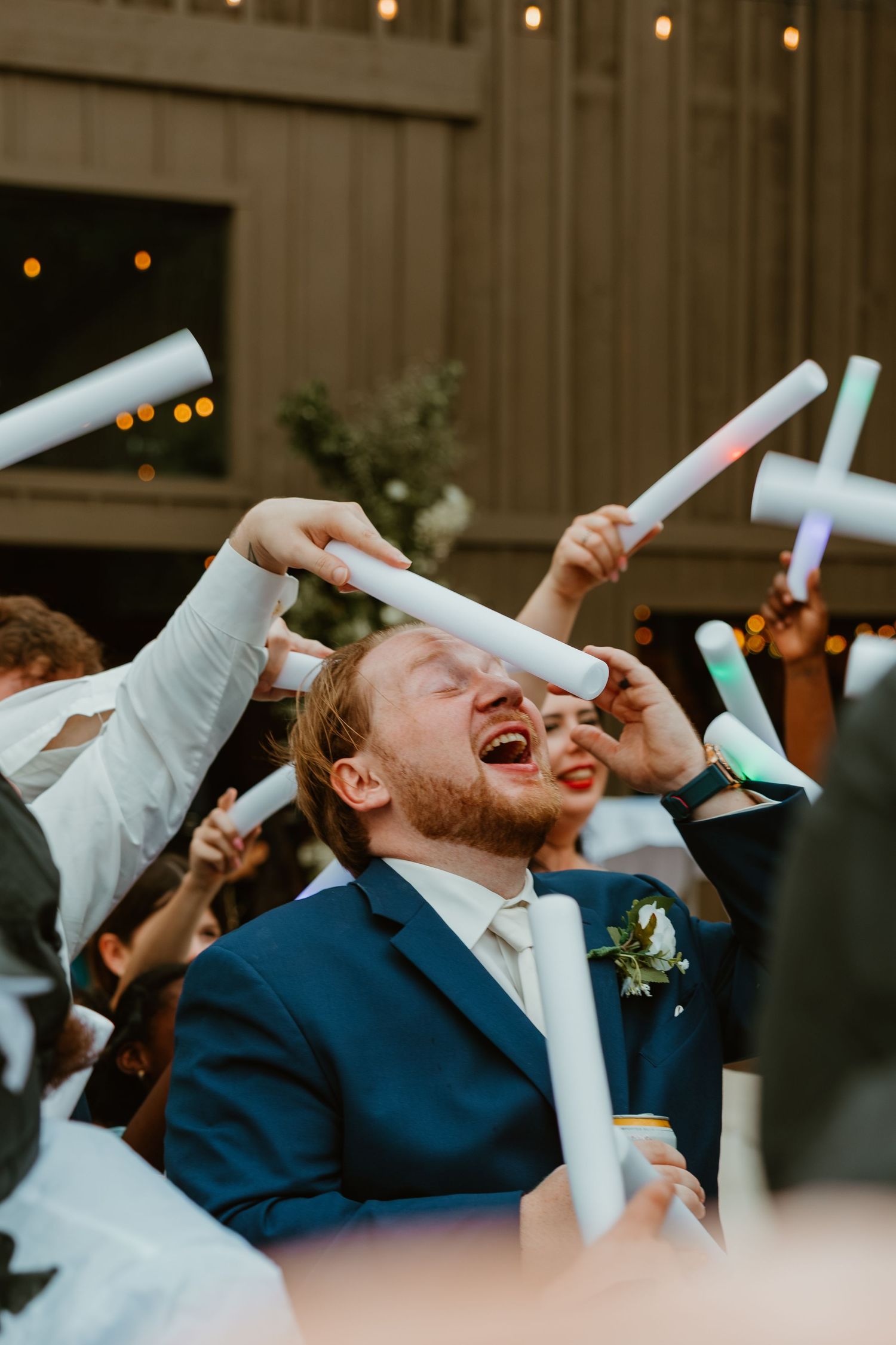 Joyful groom in navy suit celebrates with guests waving foam glow sticks at wedding reception. Northwest Indiana wedding venues.
