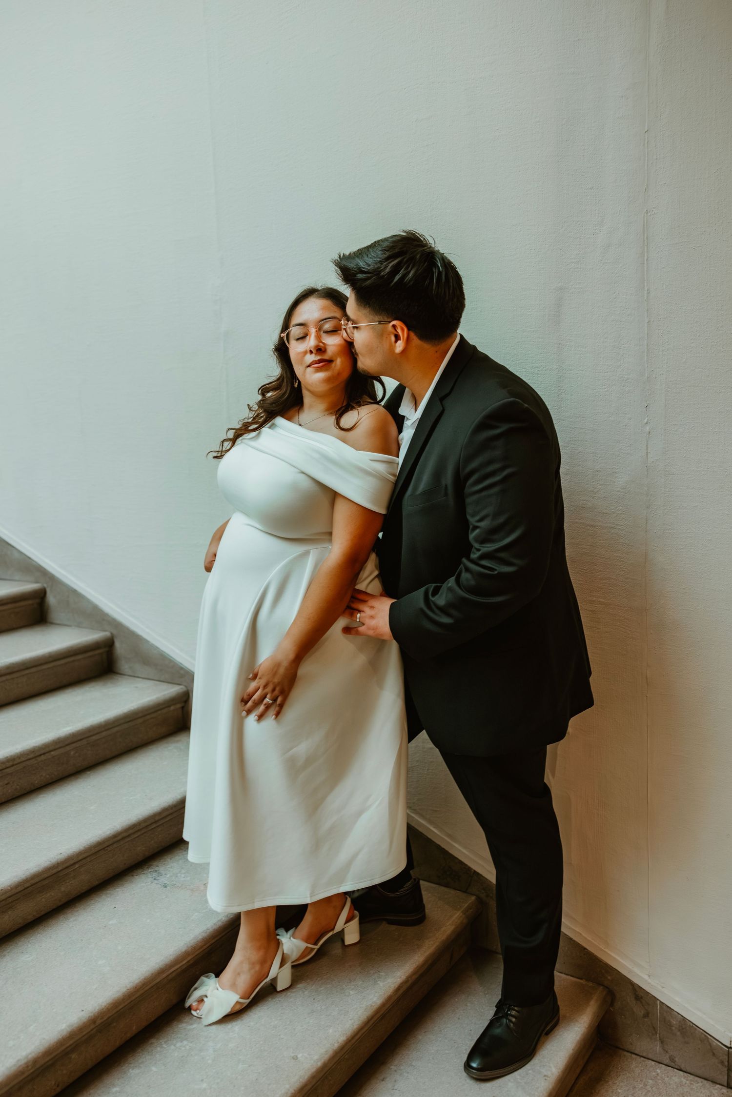 Couple in wedding attire share a romantic moment on museum stairs, groom kissing bride in white dress. Art Institute engagement photos.