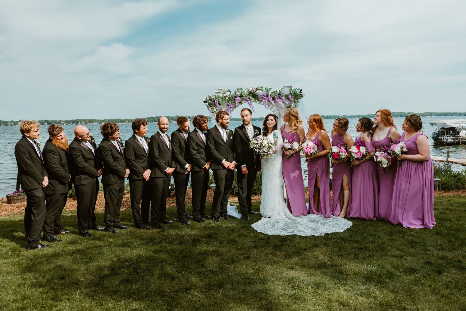 Wedding party poses by a lakeside floral arch with bridesmaids in purple and groomsmen in black.