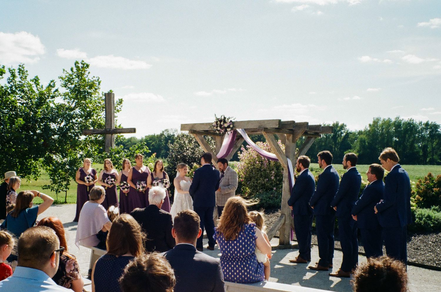 Outdoor wedding ceremony with bridal party in purple dresses and navy suits beneath a wooden arch on a sunny day.