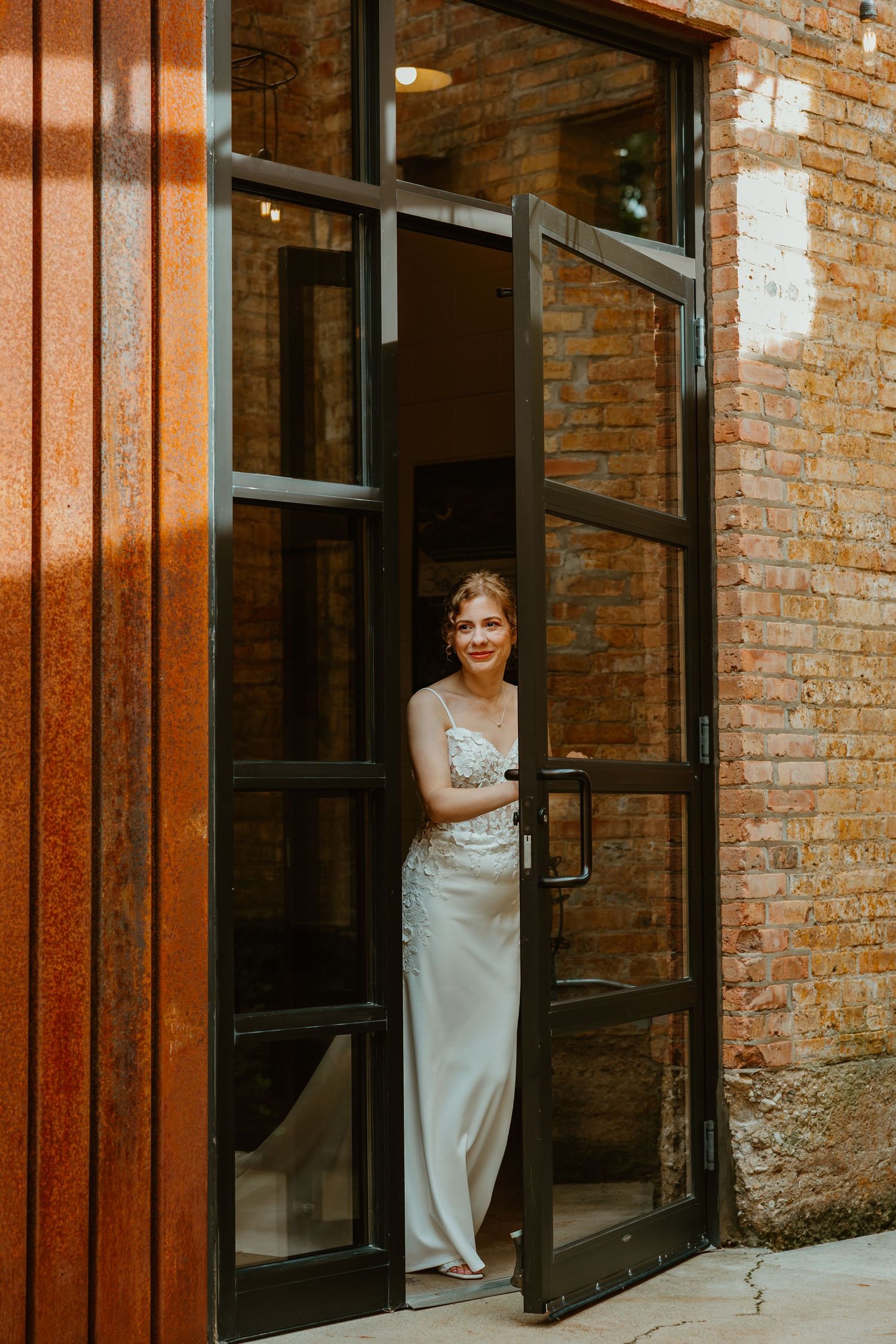 Bride in a sleek white gown standing in a black-framed glass doorway of a brick building. Chicagoland wedding venues.