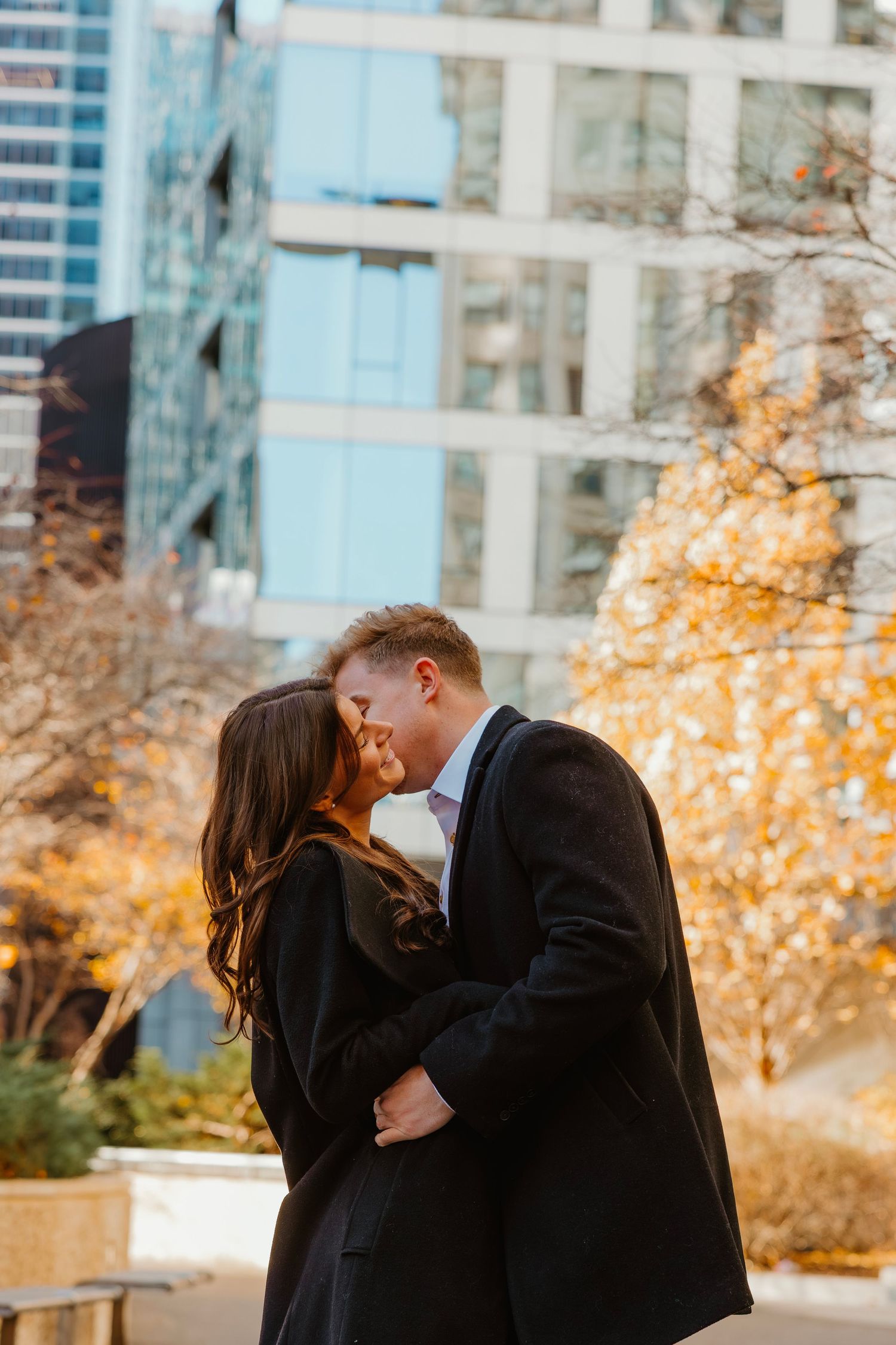 Couple sharing a tender kiss surrounded by autumn trees and modern city buildings. Outdoor Chicago engagement photos.