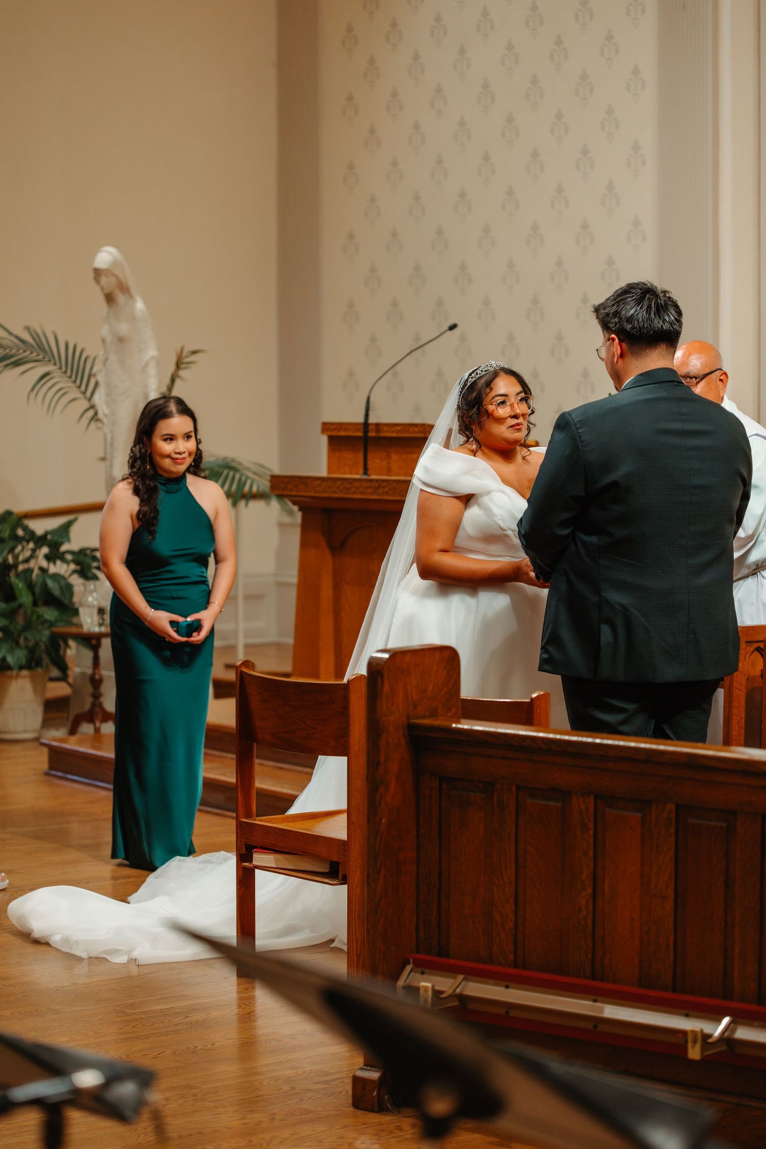 Bride smiling at groom during wedding ceremony with bridesmaid in emerald green dress. Catholic ceremony in Chicago Catholic Church.