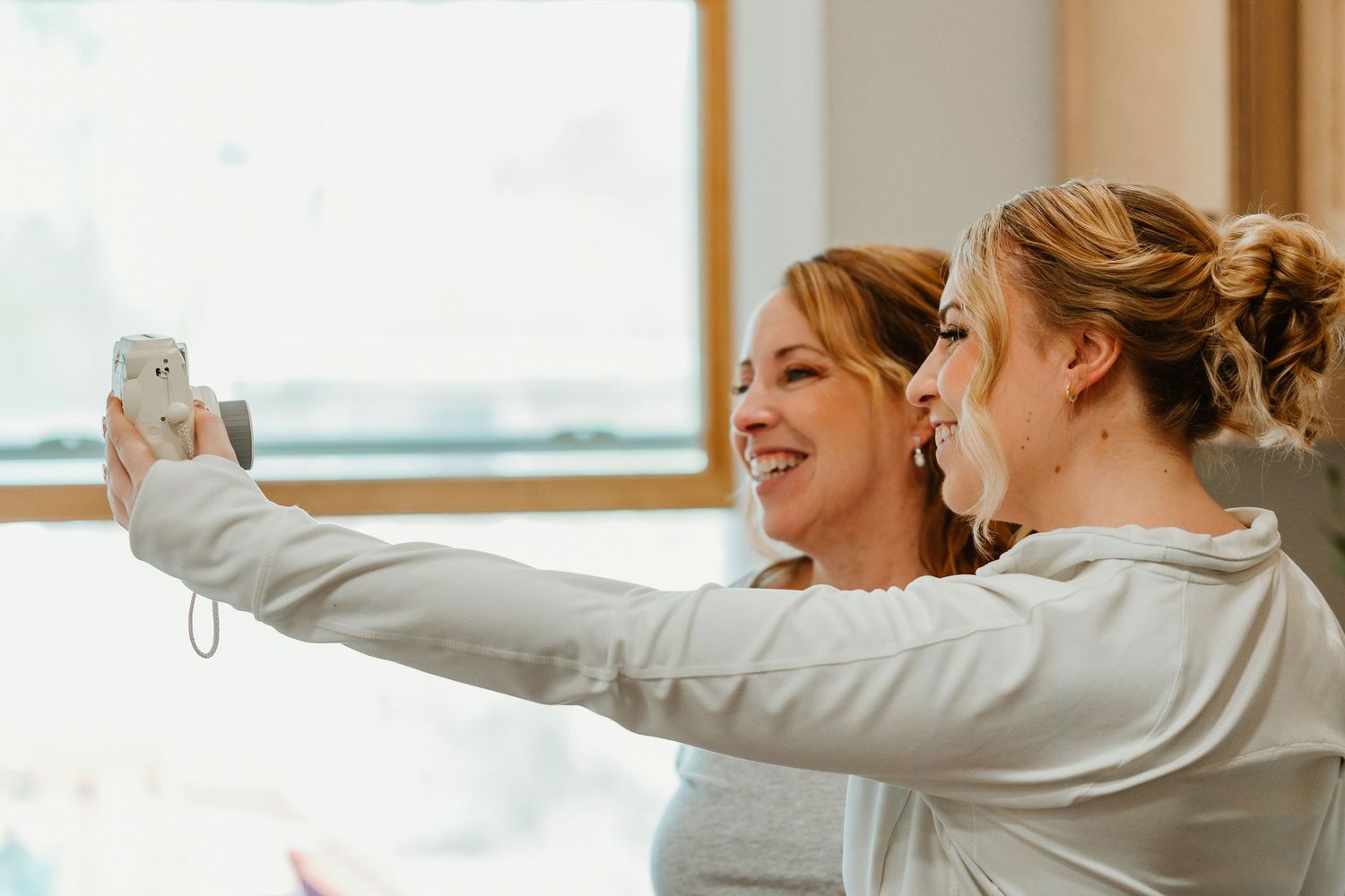 Two smiling bridesmaids in white robes taking a selfie together during wedding morning preparations. Chicago documentary style photography.