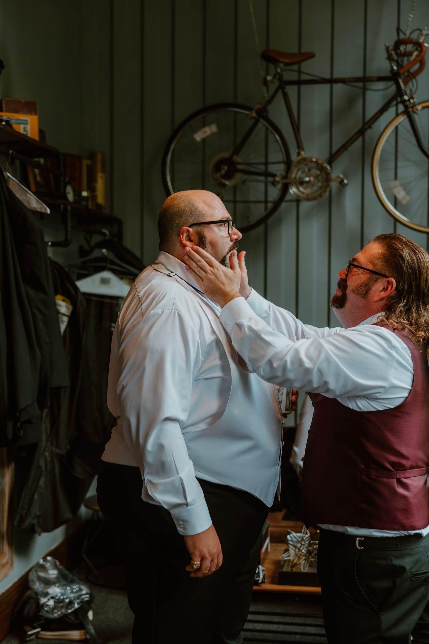 Two men in a rustic room with a bicycle on the wall, one adjusting the other's collar before a wedding. Crown Point Courthouse wedding.