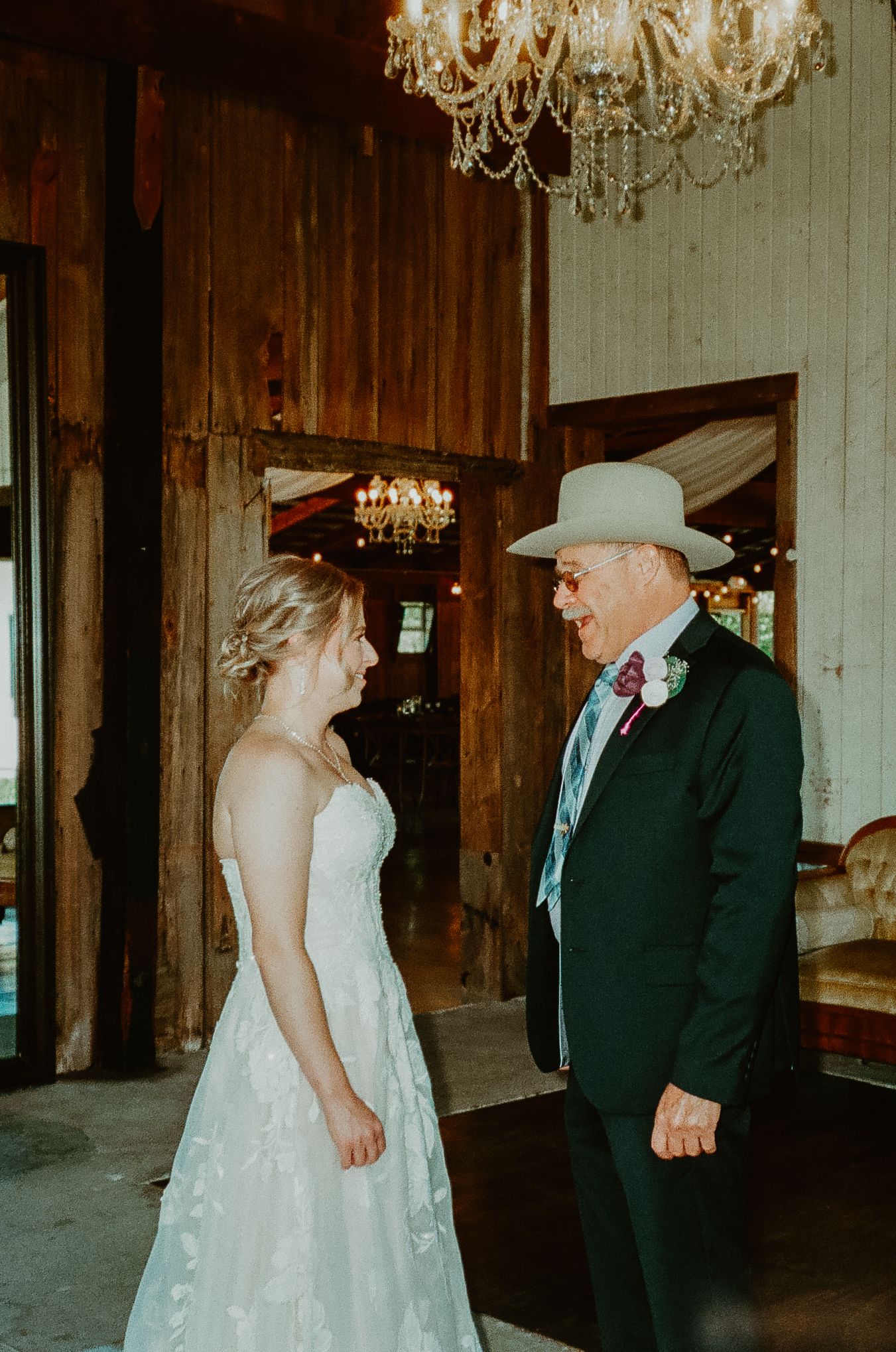 Bride in white lace gown shares a joyful first look moment with groom in cowboy hat inside rustic barn venue.
