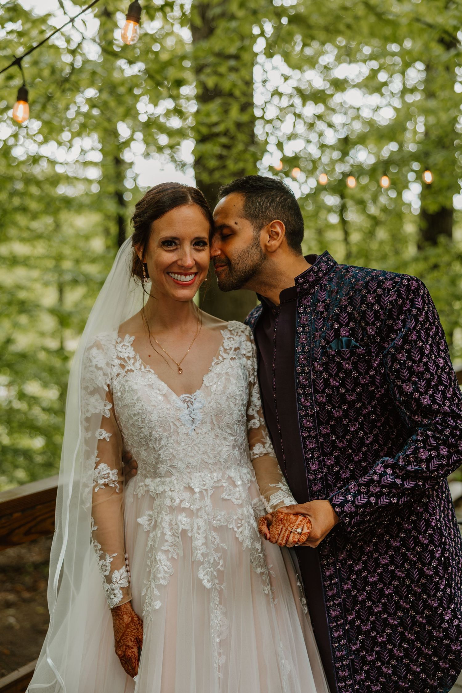 Groom kissing bride on cheek as she smiles in a lace wedding gown with veil outdoors. Documentary style wedding photos.