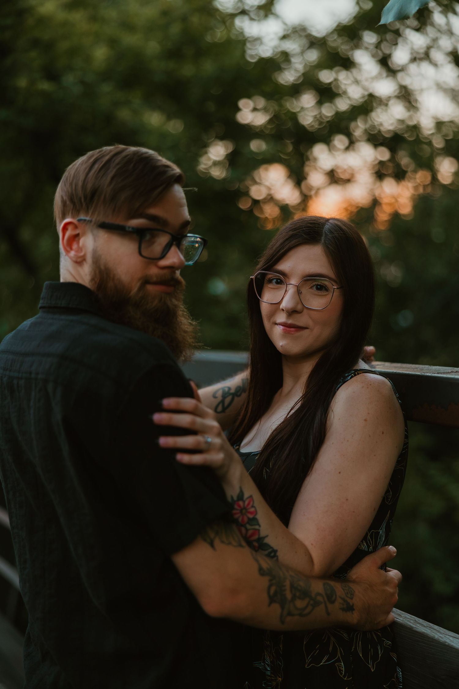 Tattooed couple embracing on a bridge at sunset, both wearing glasses, woman smiling at camera. Chicagoland engagement photos.