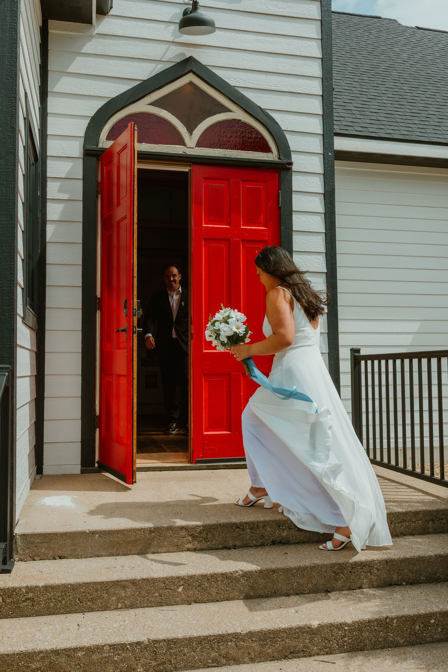 Bride in white gown carrying bouquet walks up steps toward groom at red church doors. Michigan elopement.