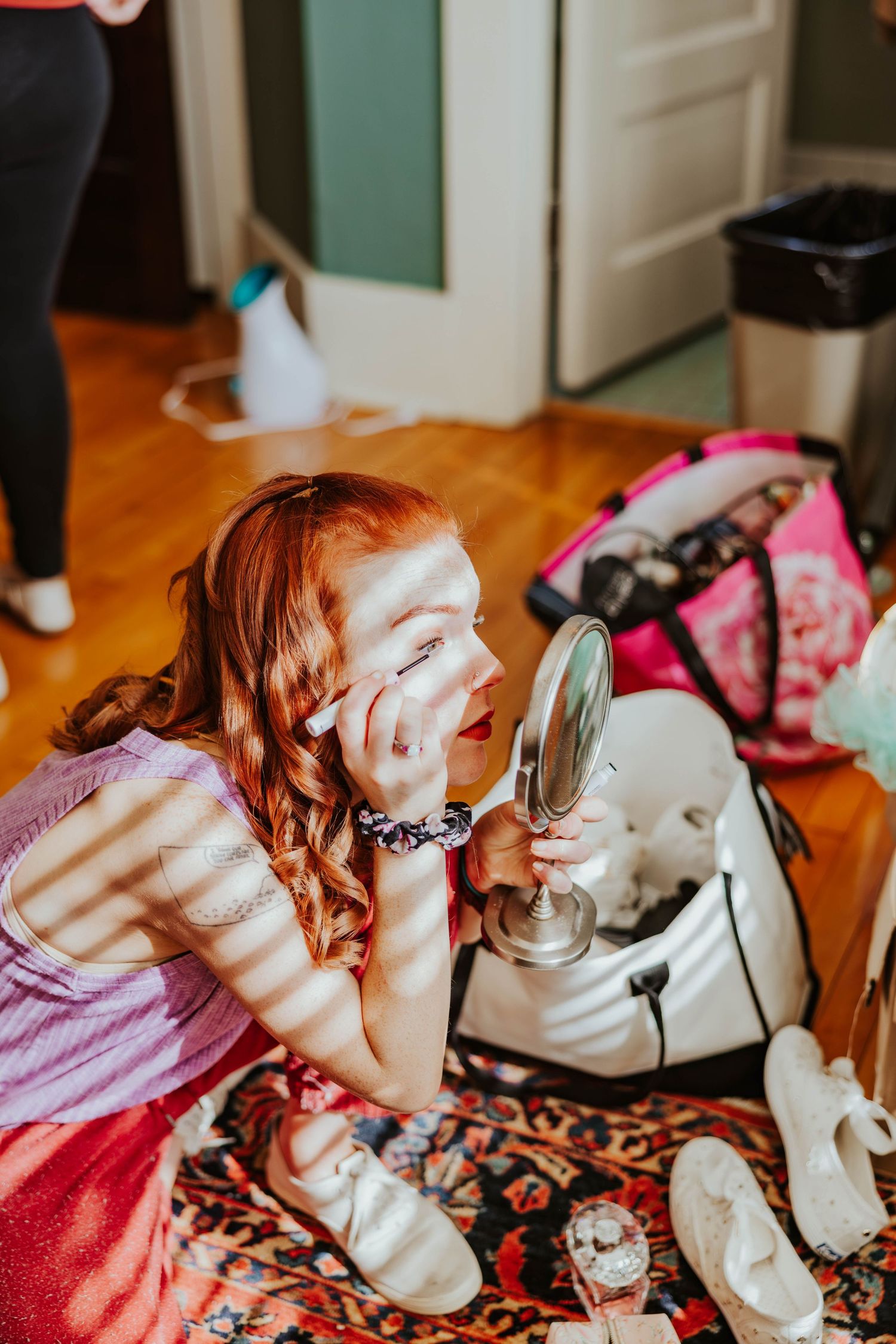 Red-haired bridesmaid applies eye makeup using hand mirror while sitting on floor during wedding prep.