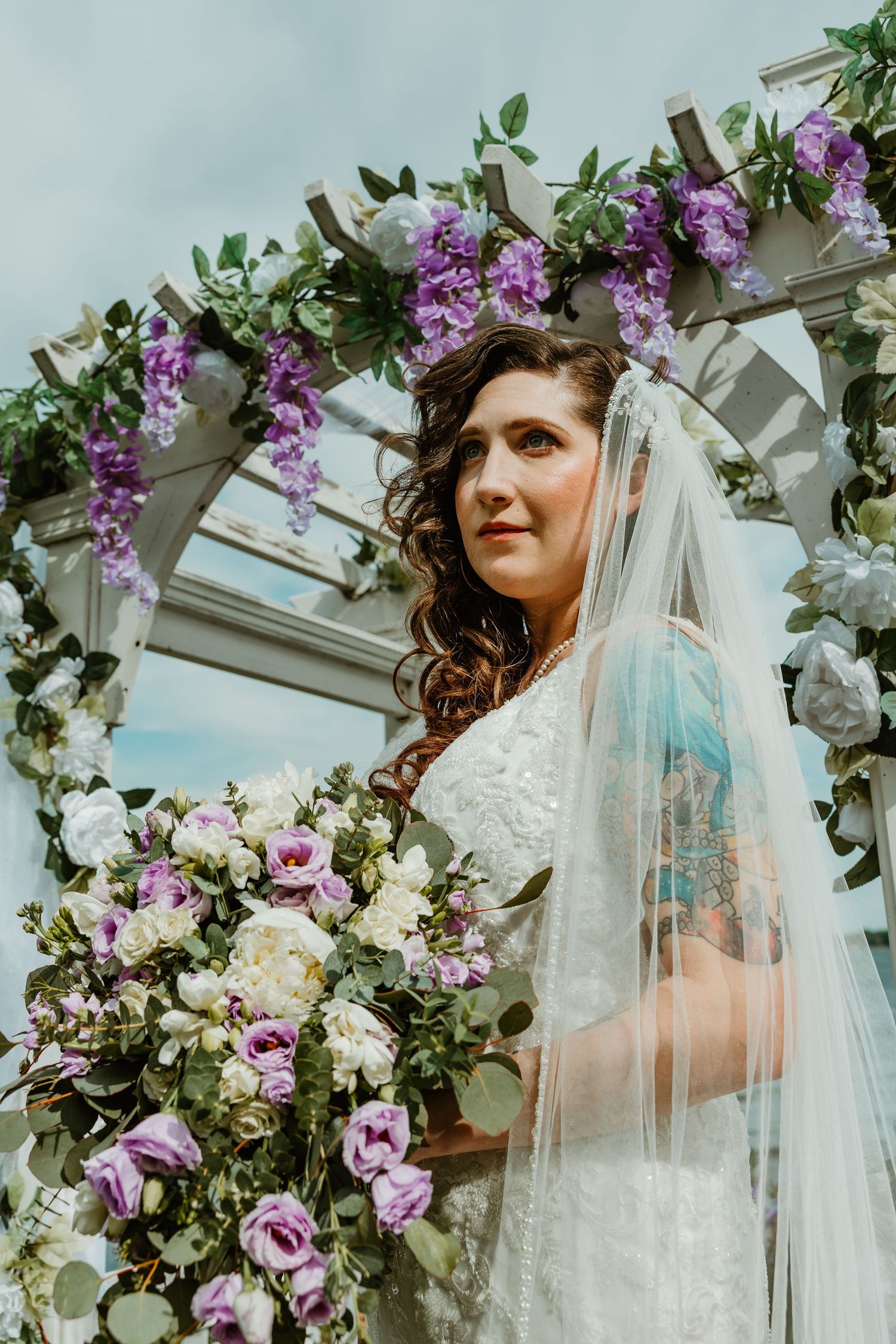 Bride in lace gown and veil poses under floral arch adorned with purple wisteria and white roses.