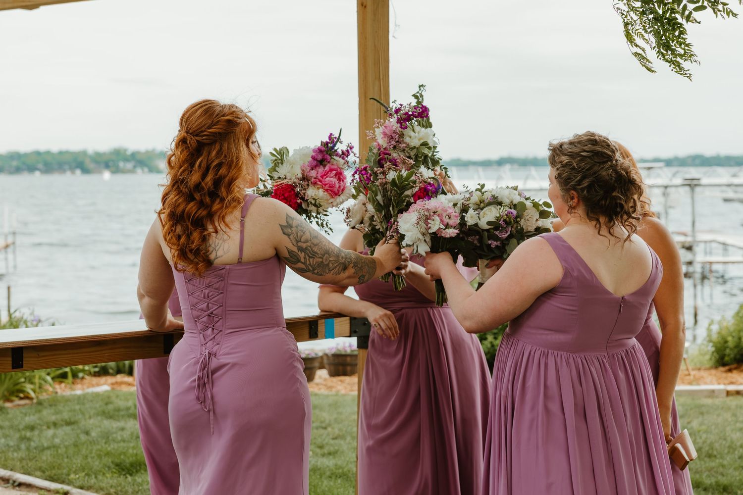 Bridesmaids in dusty rose gowns hold colorful bouquets near a wooden arch by the water.