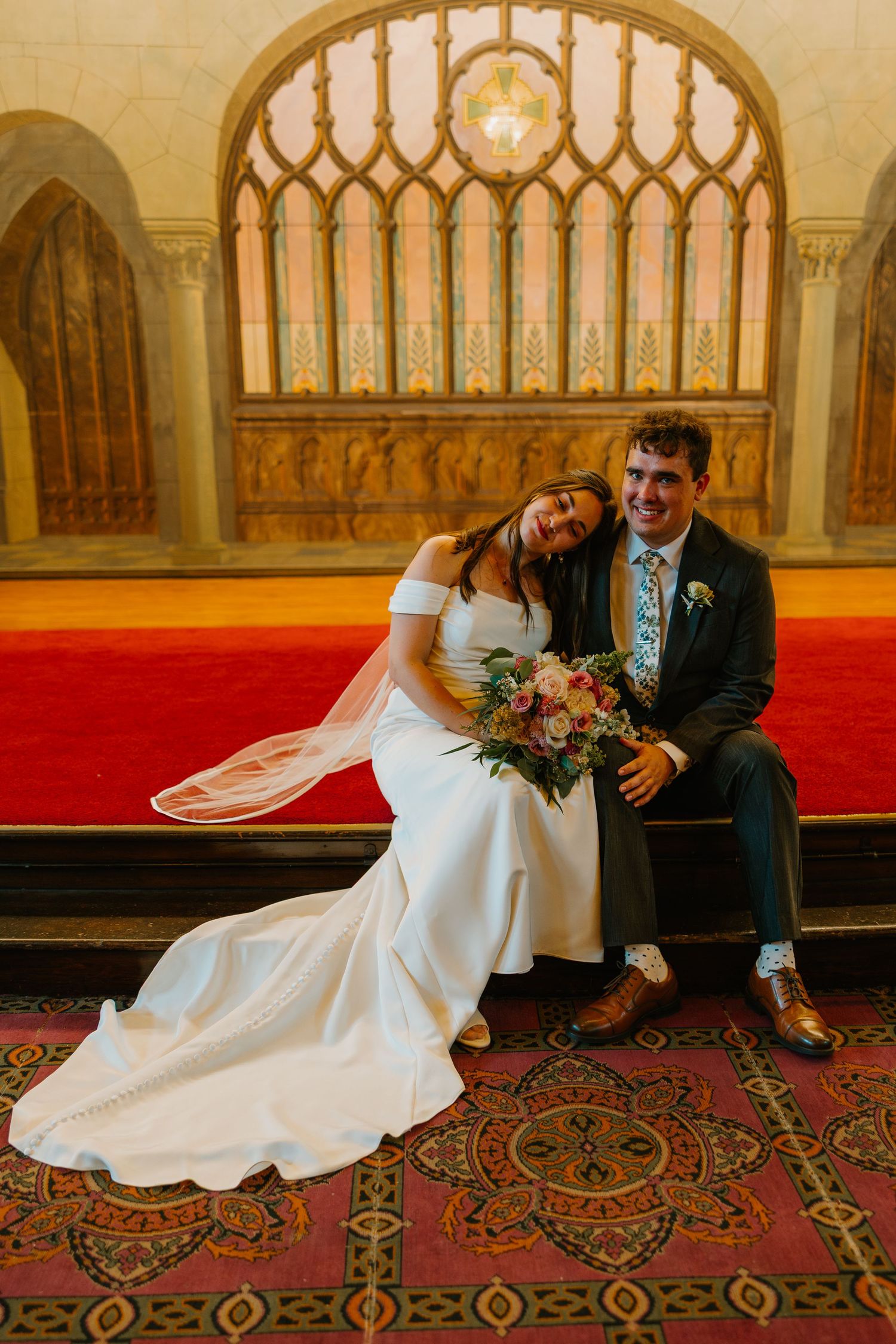Bride and groom sitting together on red carpet bench in front of Gothic arched stained glass window.