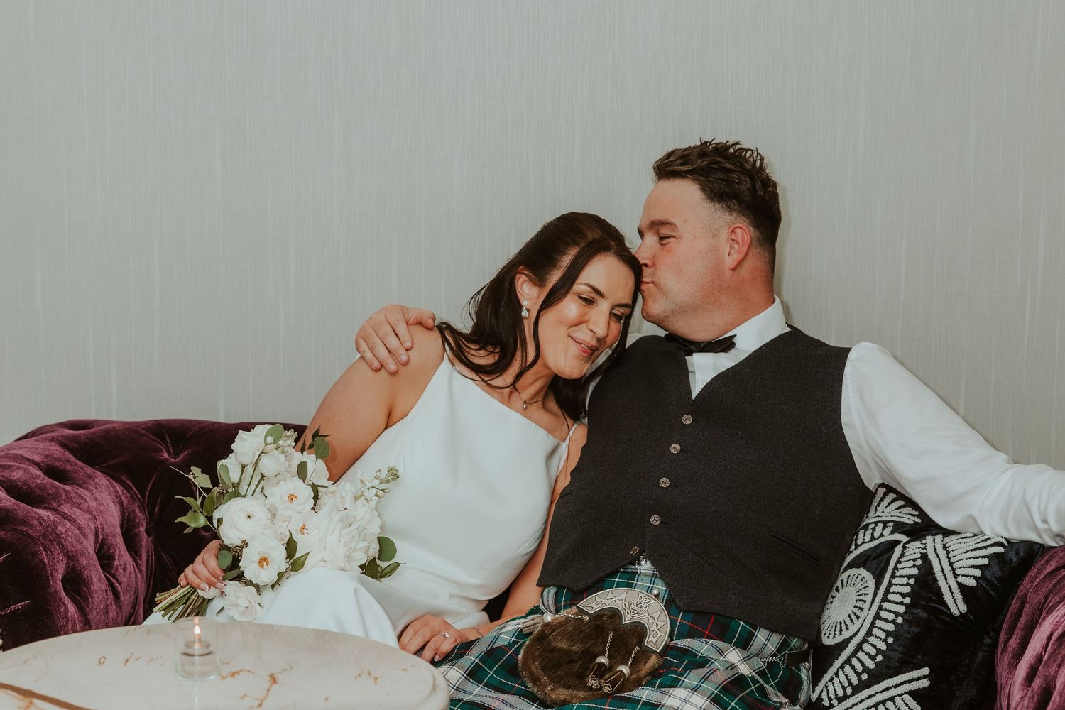 Bride and groom sharing a tender moment on a velvet sofa, groom kissing her cheek with bouquet nearby. Chicago Thompson Hotel wedding.